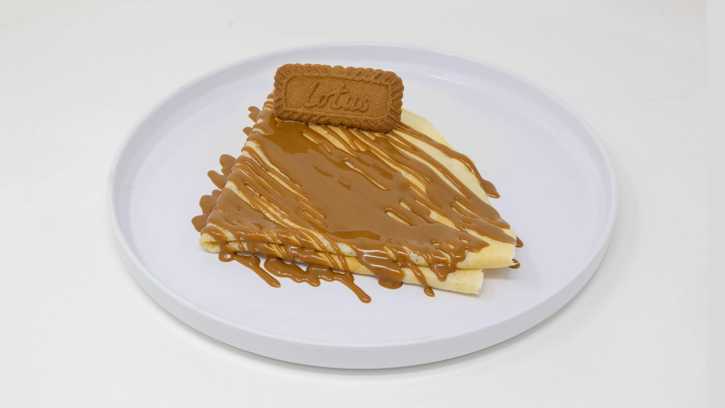 A slice of crepe cake drizzled with caramel sauce, topped with a Lotus biscuit on a white plate.