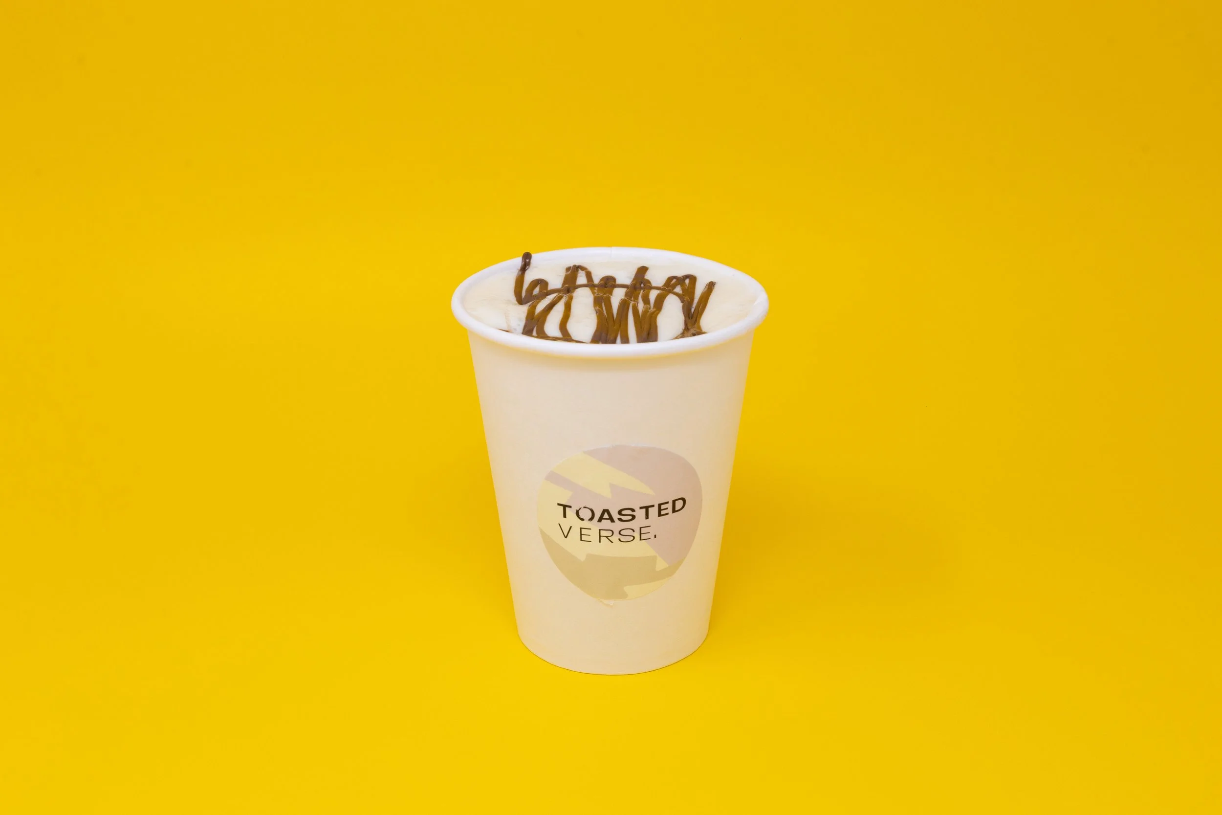A white cup with a beige label reading "Toasted Verse" contains a beverage topped with whipped cream and chocolate drizzle, set against a bright yellow background.