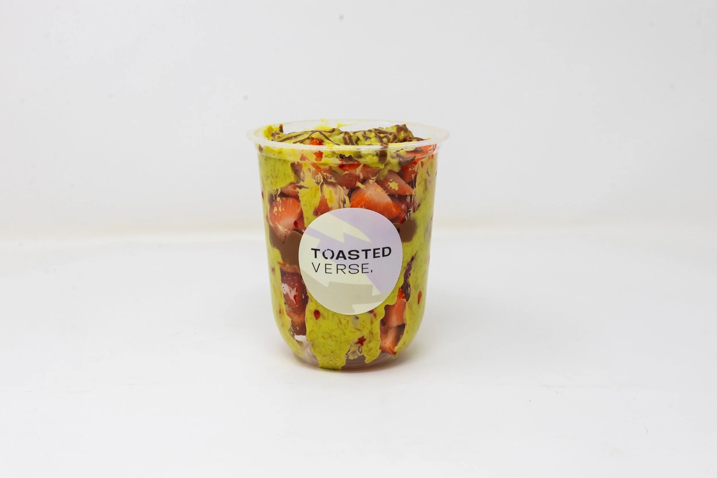 Clear plastic cup with green and red fruit pieces, topped with a drizzle of chocolate sauce, labeled 'Toasted Verse'.