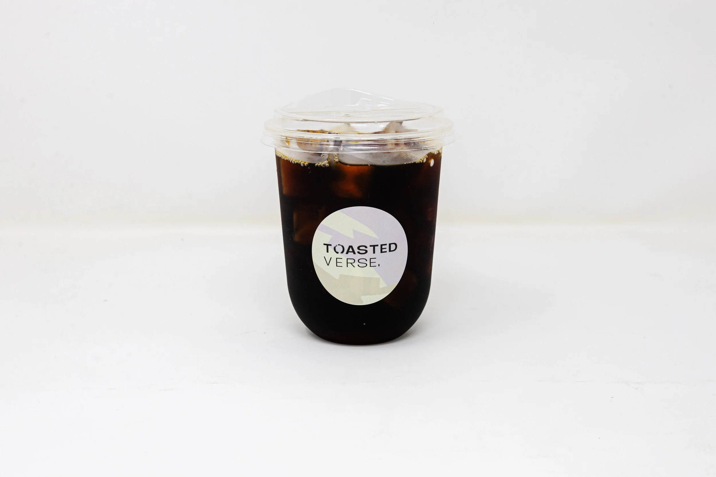 Plastic cup of iced dark coffee with a clear lid, label reading "Toasted Verse," placed on a plain white surface with a white background.