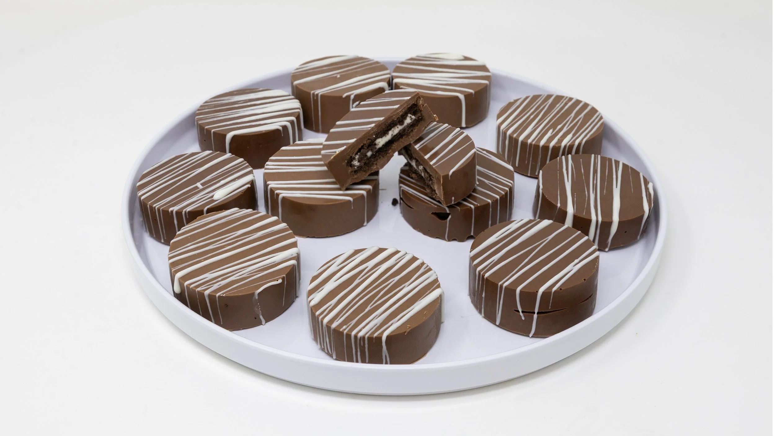 A white tray filled with round chocolate candies topped with white chocolate drizzle, with one broken in half revealing a white filling inside.