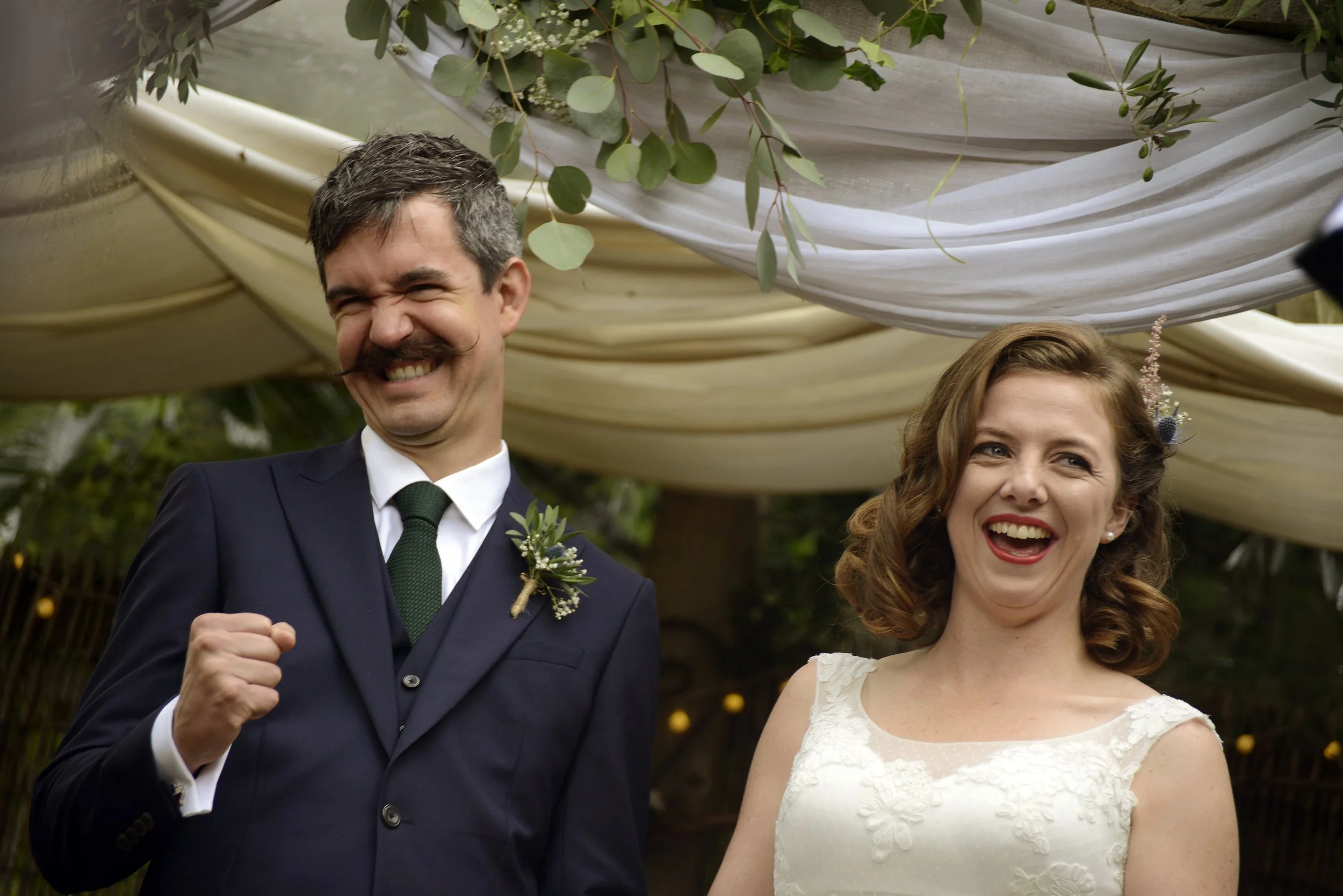 A joyful groom in a dark suit and tie and a smiling bride in a white lace wedding dress at their outdoor wedding ceremony, decorated with draped fabric and greenery.