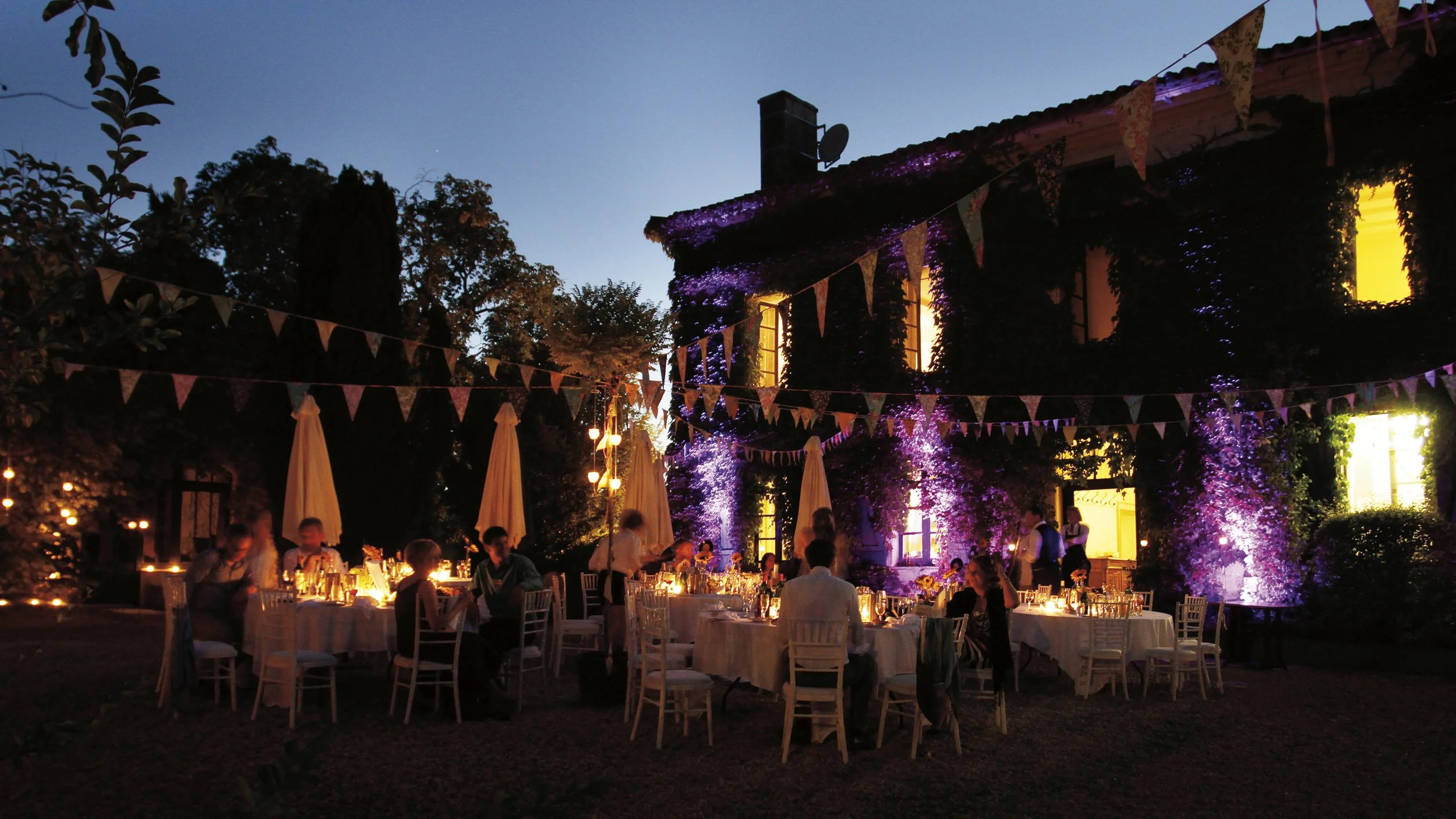 An outdoor evening dinner party with multiple tables illuminated by candles, surrounded by people, set against a building decorated with purple and yellow lighting, with bunting flags hanging overhead and trees in the background.