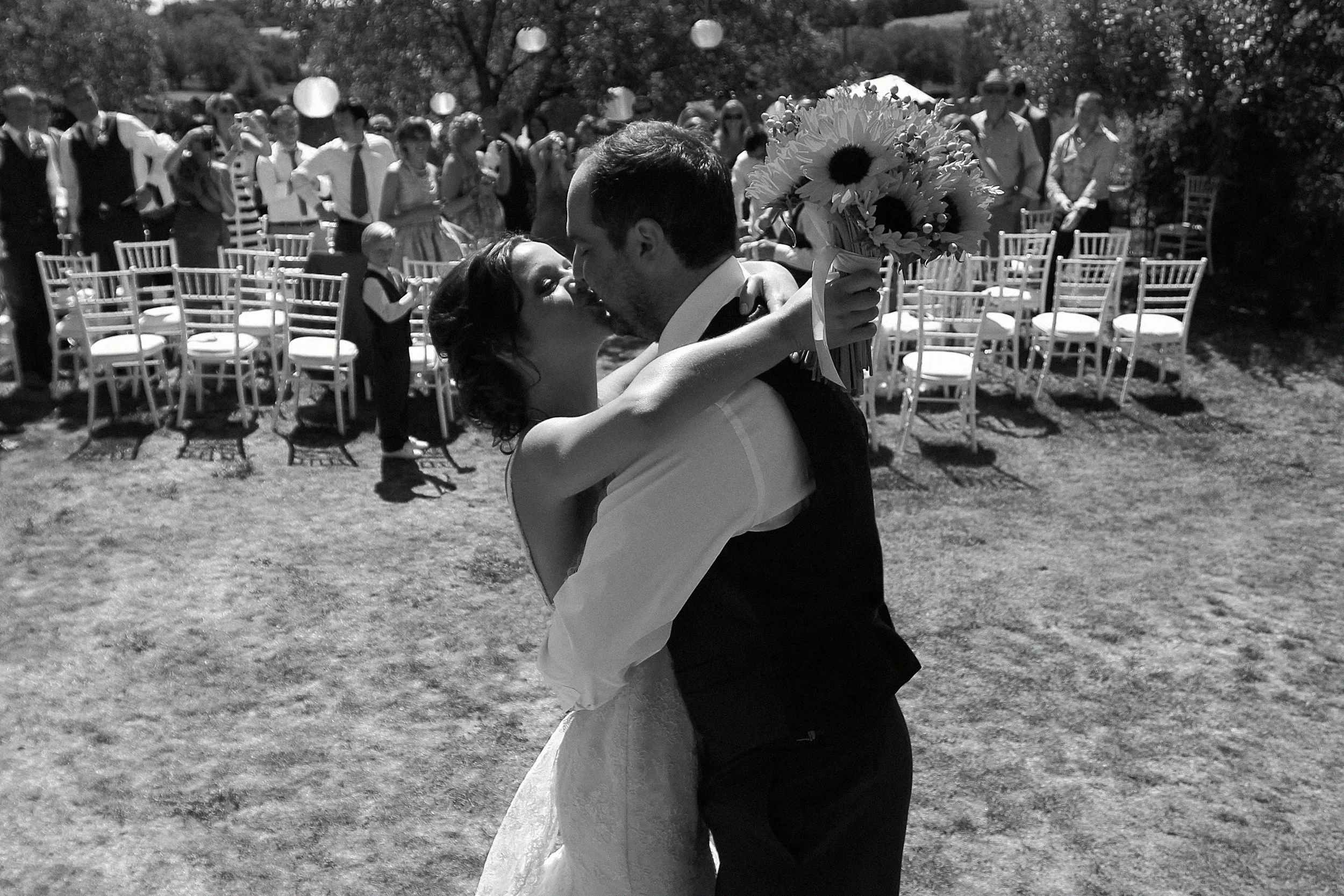 A bride and groom share a kiss outdoors at their wedding, surrounded by seated guests and standing on a grassy area with chairs and trees in the background.