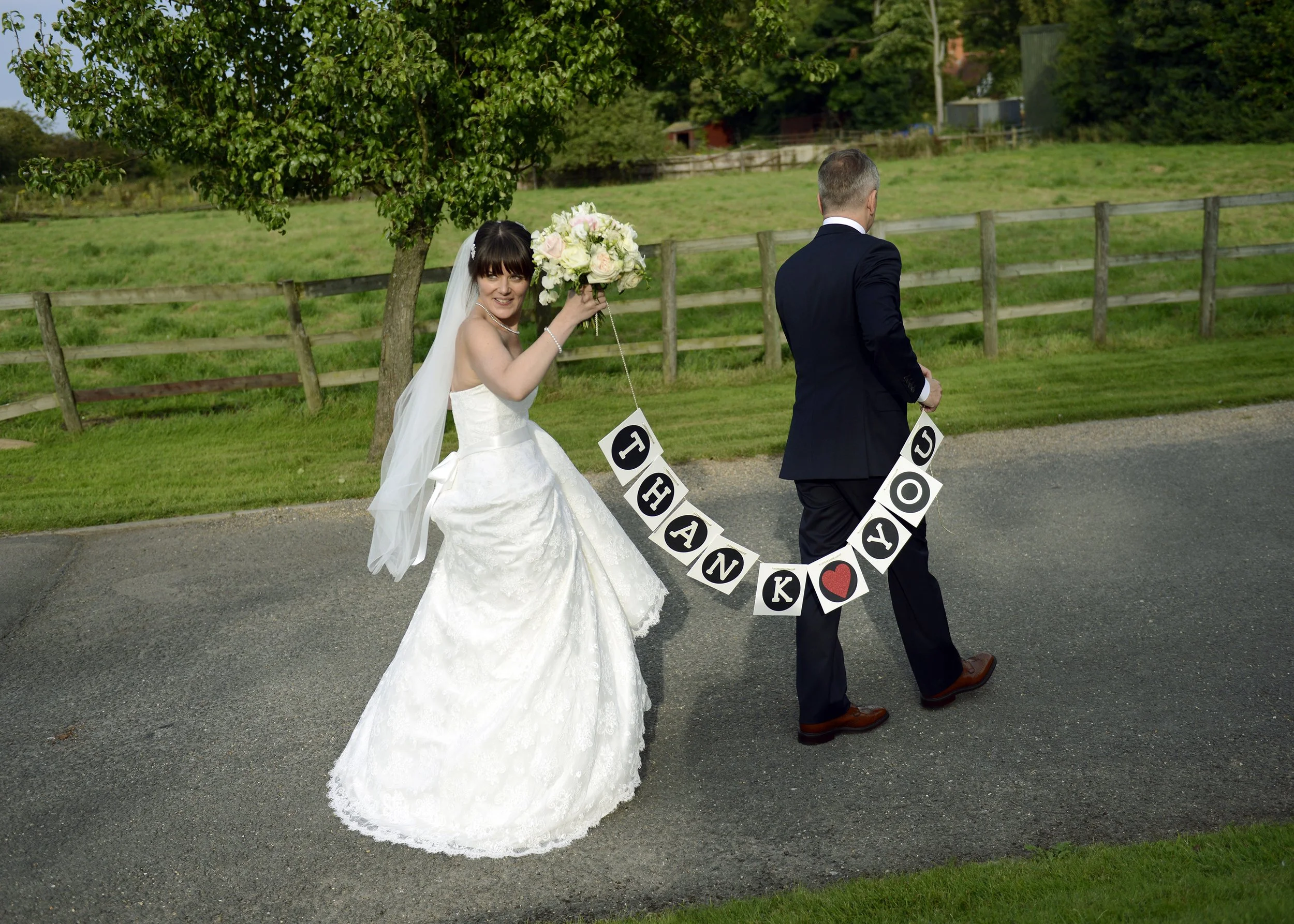 A newlywed couple, walking away from the camera on a paved path in a lush green outdoor setting. The bride, in a white wedding dress, is smiling and holding a bouquet of flowers in one hand and a sign that says "THANK YOU" with a red heart in the oth