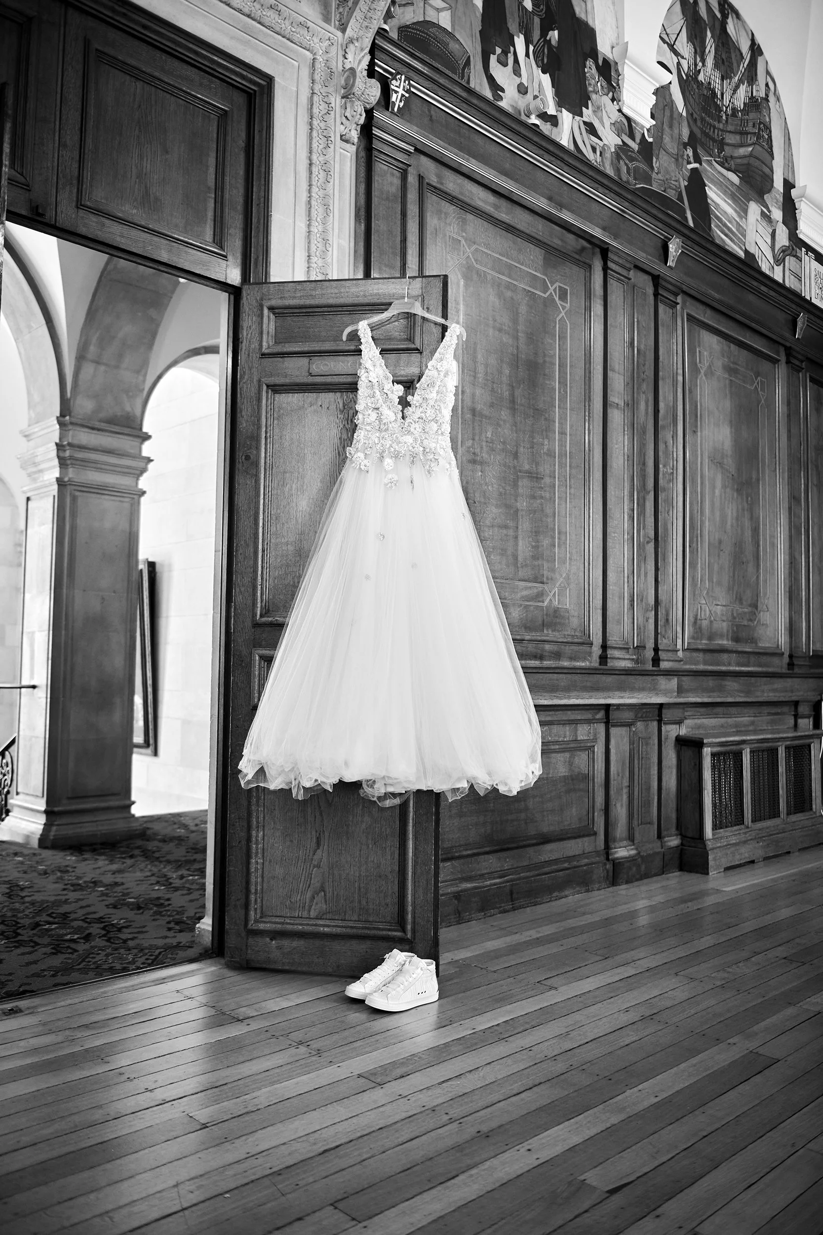 A wedding dress hanging on a hanger inside a wooden-paneled room with a pair of white sneakers on the floor below.