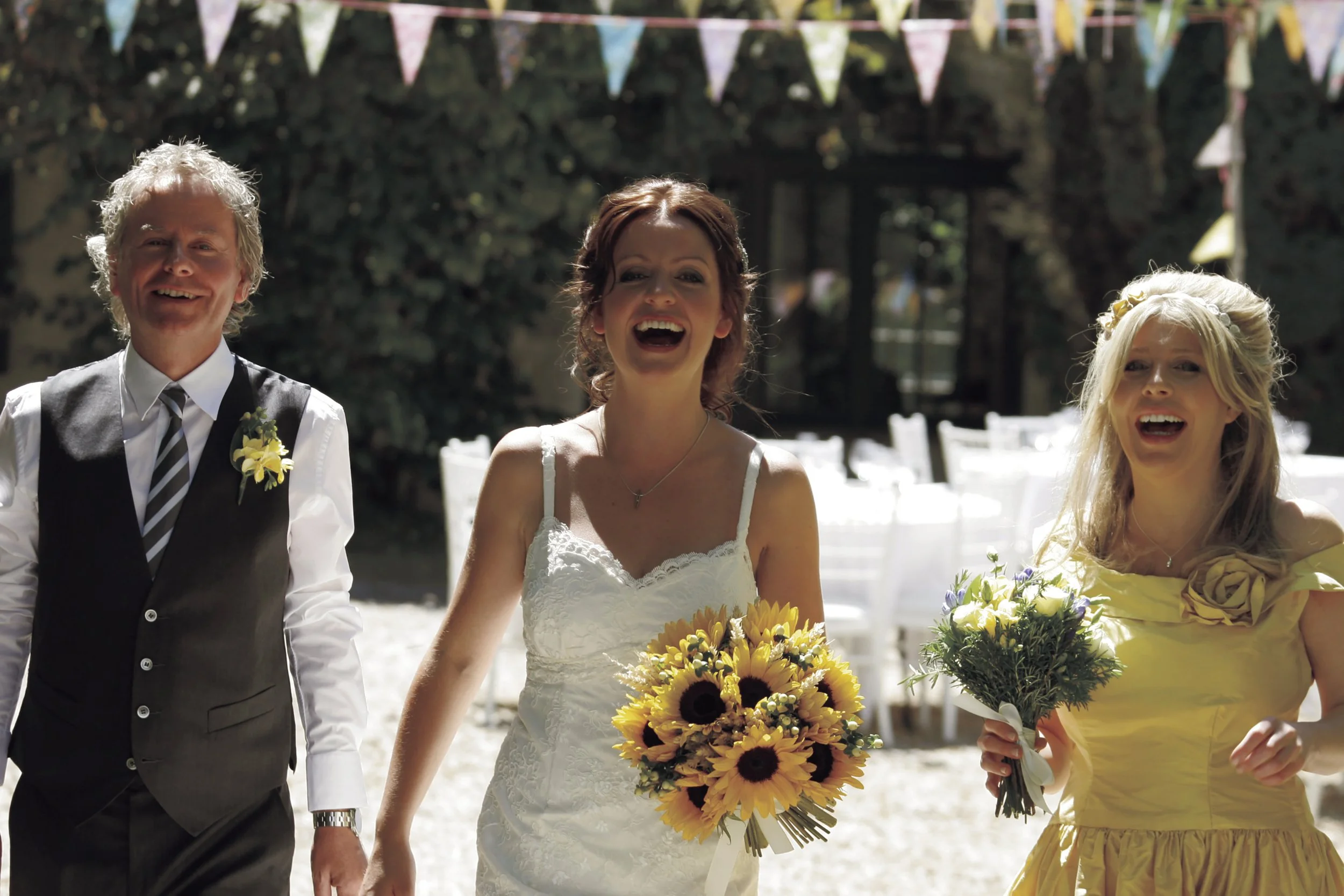 A bride in a white dress holding a bouquet of sunflowers, walking with two women and a man, all smiling, at an outdoor wedding ceremony with bunting and trees in the background.