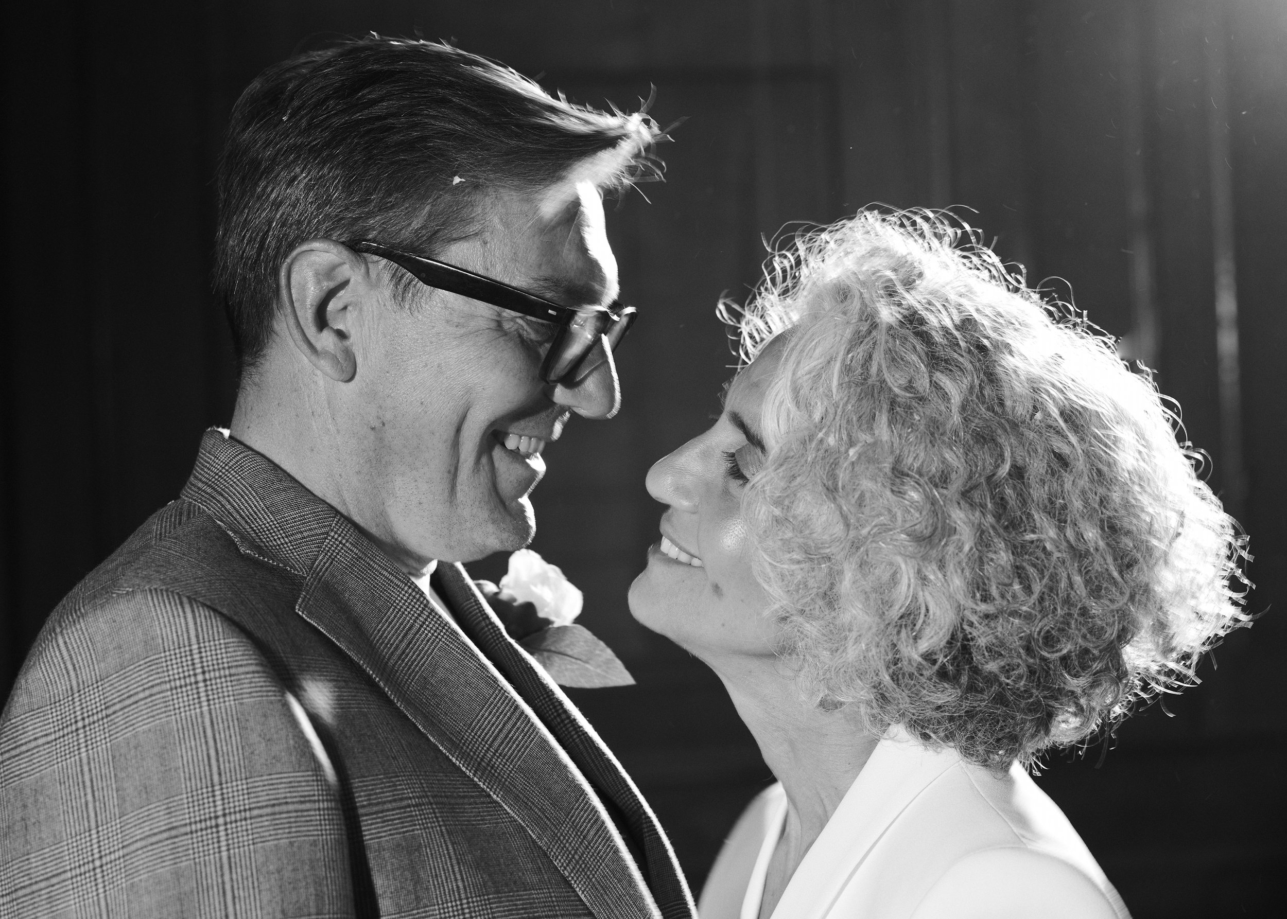 A man and a woman face each other smiling close together. The man is wearing glasses and a checked jacket, and the woman has curly hair and is wearing a light-colored outfit. The photo is in black and white.