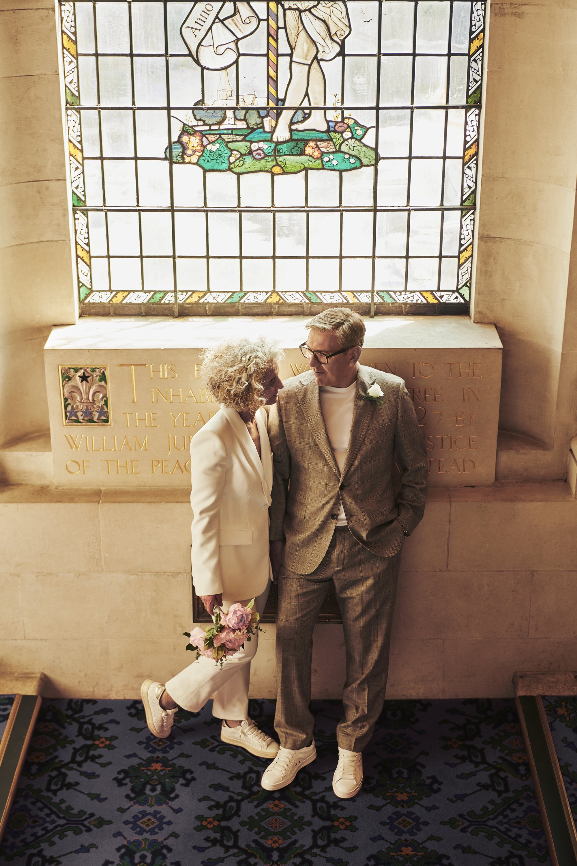 A woman with curly gray hair in a white suit holding a bouquet of pink flowers, and a man with short blonde hair in a gray plaid suit, standing close and looking at each other inside a church with a stained glass window behind them.