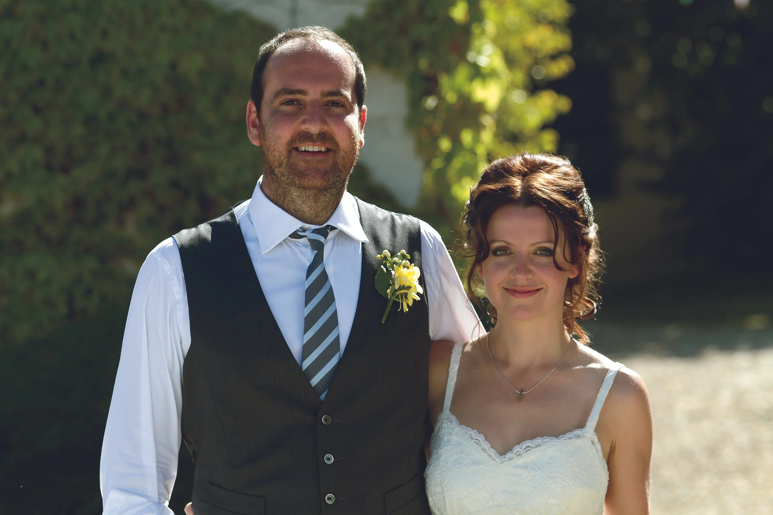 A man and woman dressed in wedding attire standing outdoors by a body of water, smiling at the camera.