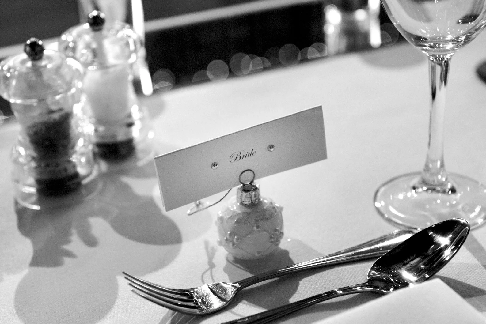 Wedding reception table setting with a 'Bride' name card, a wedding ring, a white ornament, a spoon, a fork, a wine glass, and salt and pepper shakers in black and white.