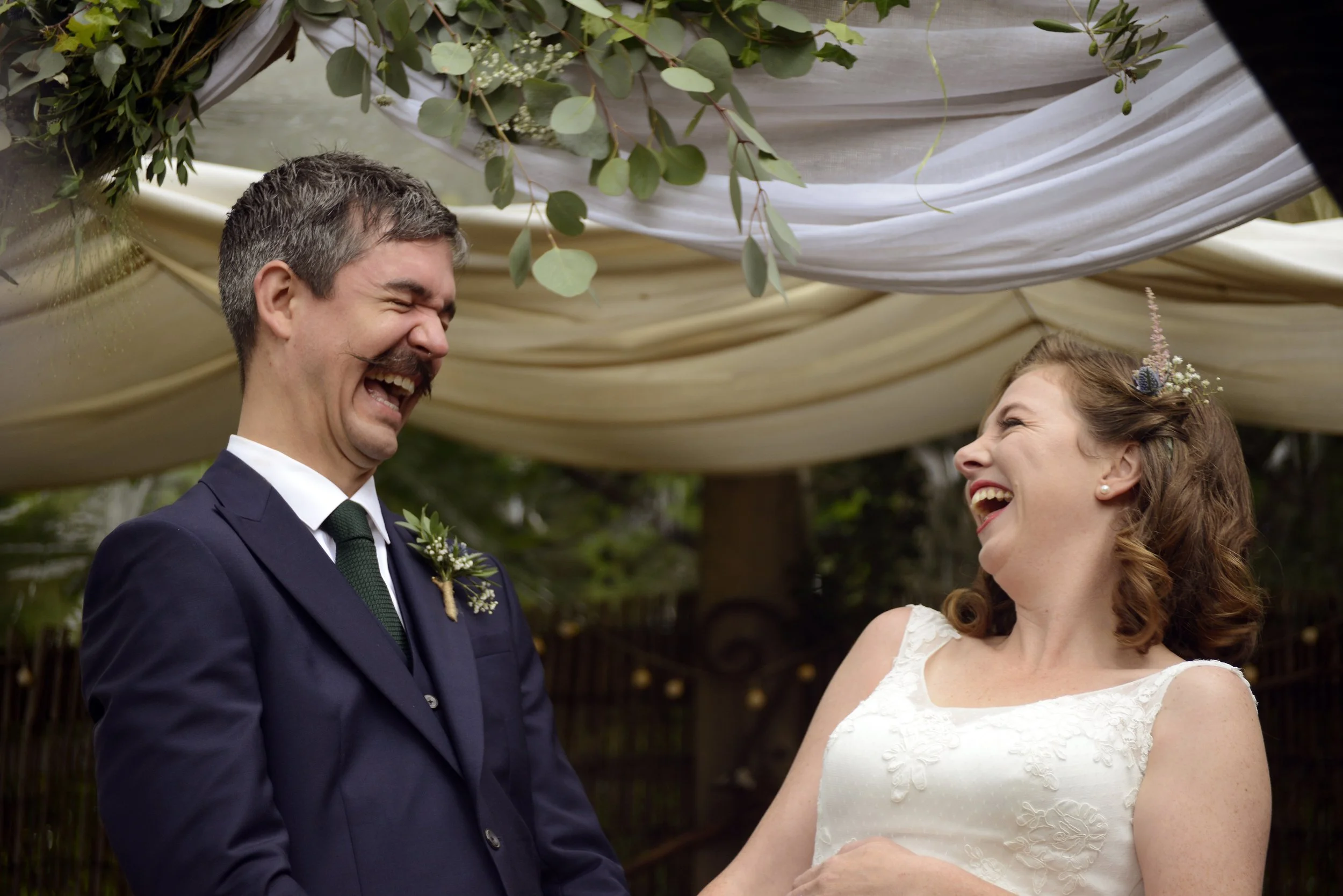 A bride and groom laughing together at their outdoor wedding ceremony, with white drapery and green foliage above them.