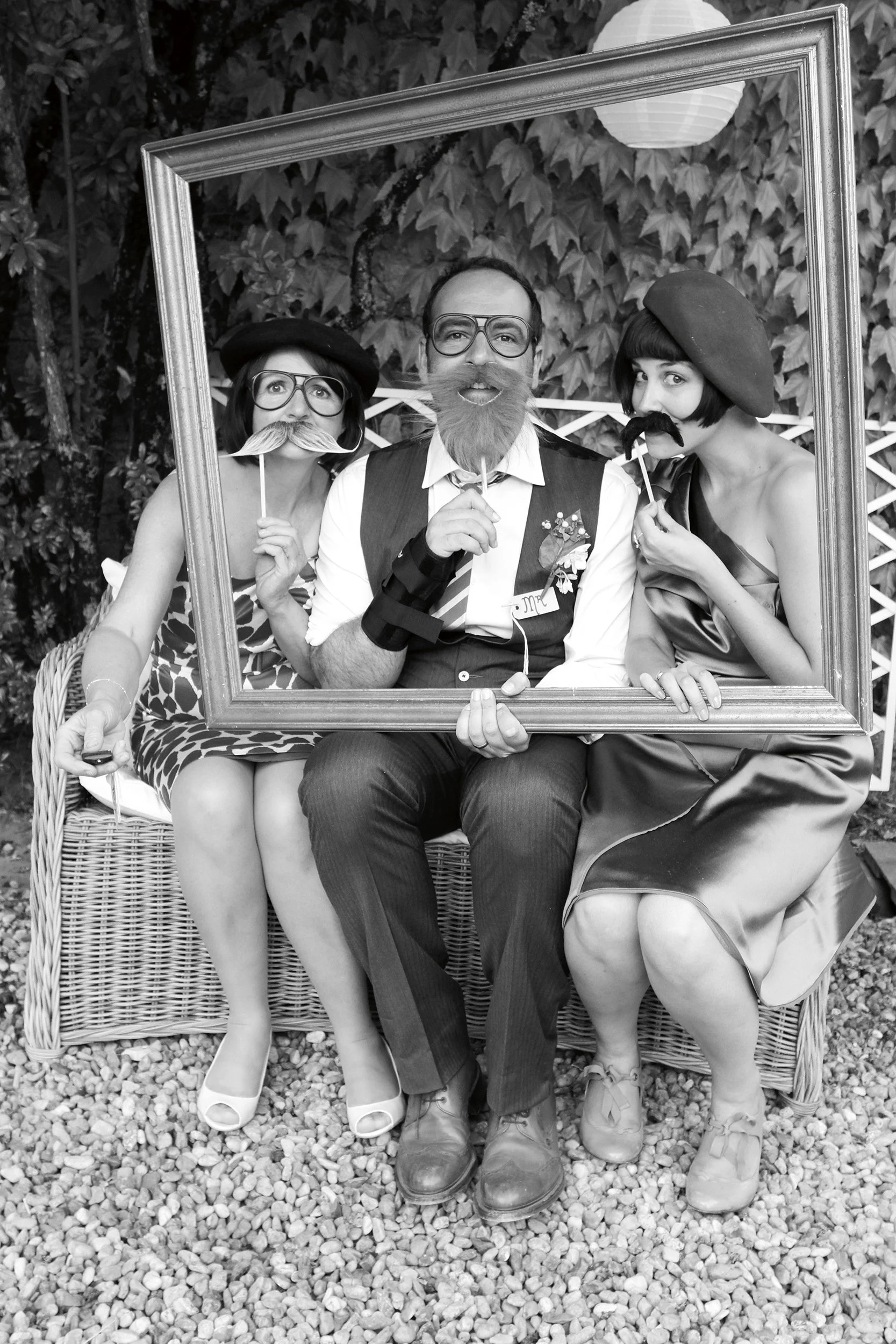 Three people sitting on a wicker bench behind a large rectangular frame, dressed in vintage clothing and holding fake mustaches and glasses, with a leafy background.