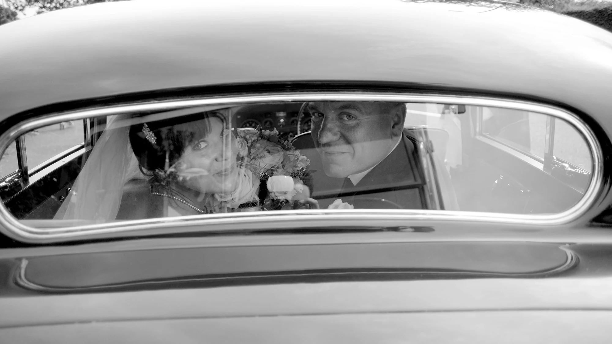 Black and white photograph of a bride and groom inside a classic car, seen through the rear window, looking at the camera. The bride is holding a bouquet, wearing a veil, and the groom is smiling.