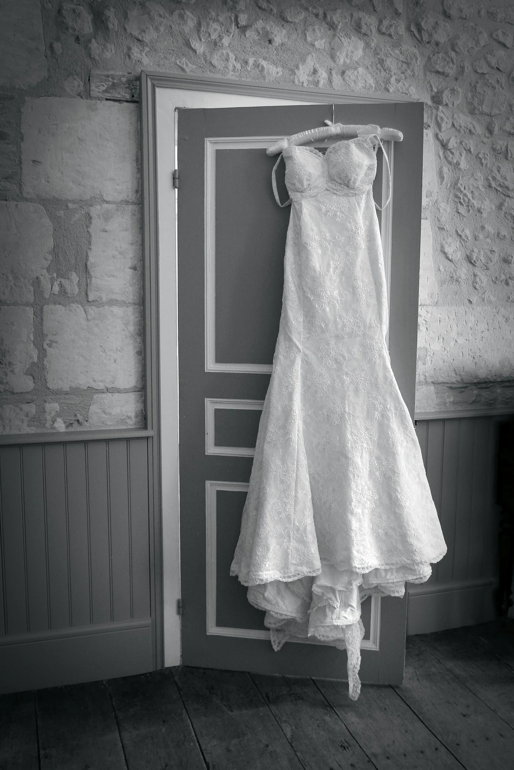 A white wedding dress hanging on a wooden hanger on a grey door, with a stone wall background.