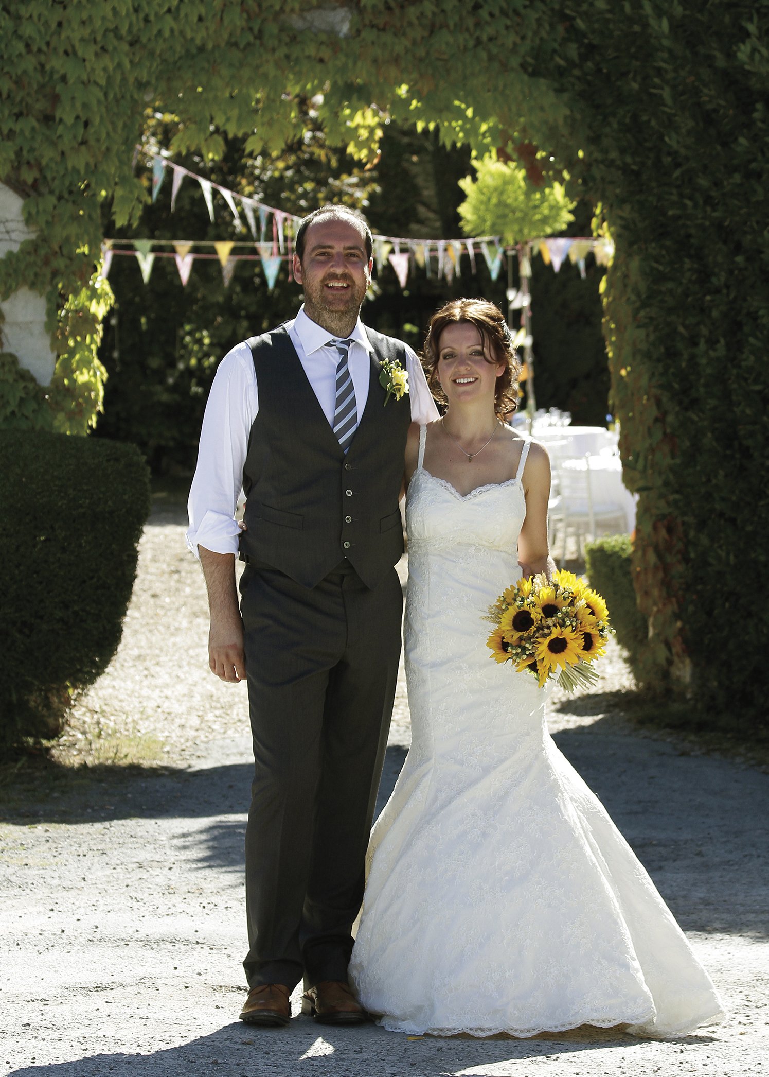 A newlywed couple standing outdoors under a decorative arch, smiling, with the bride holding a bouquet of sunflowers and the groom wearing a suit and striped tie.