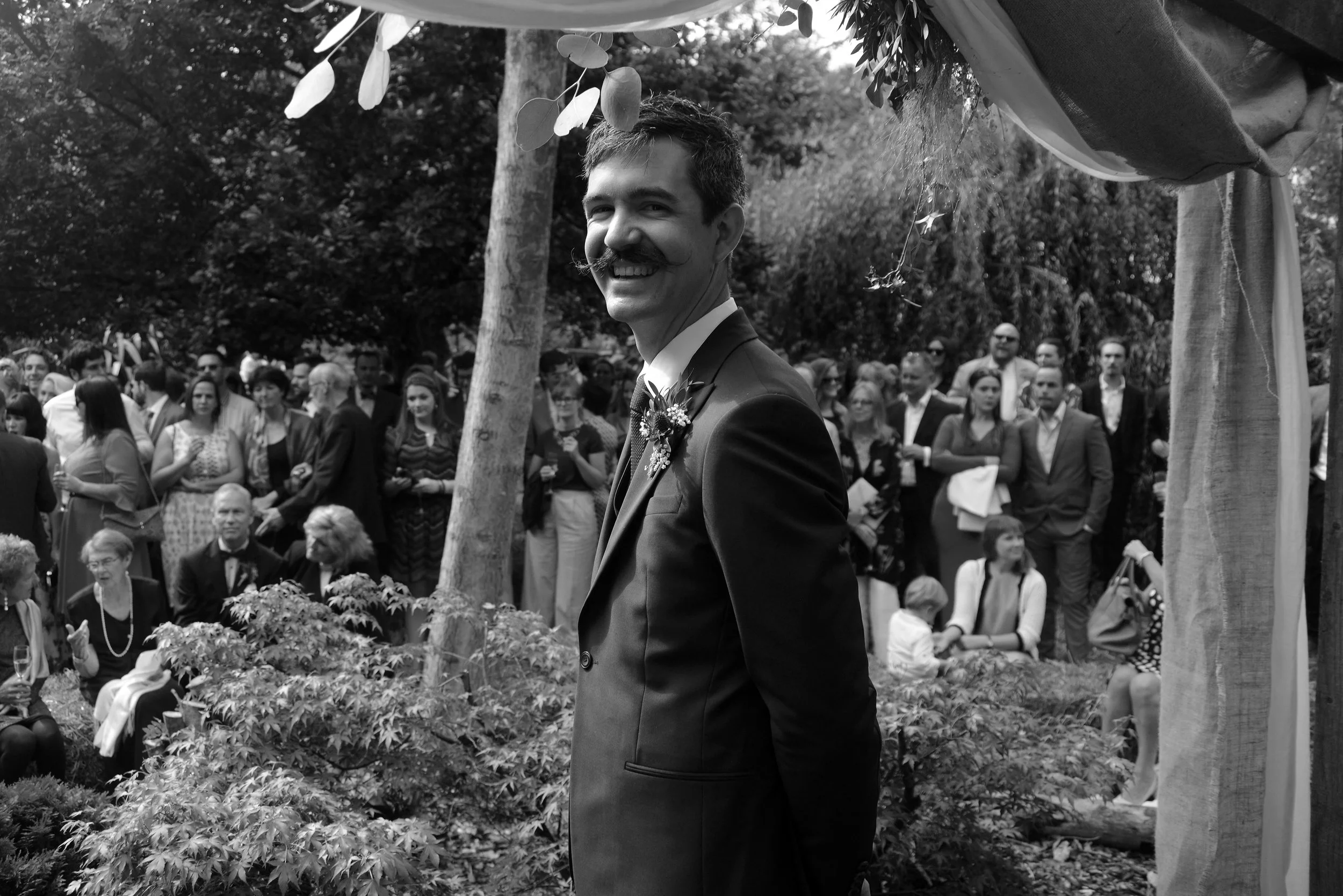 A smiling man with a mustache dressed in a suit at an outdoor event, with a large group of people gathered around and trees in the background.