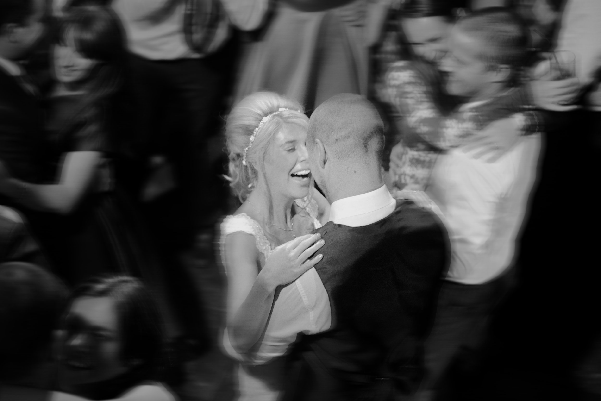 A woman in a wedding dress and a man in a suit dance closely, smiling, with other people in the background at a celebration or wedding.