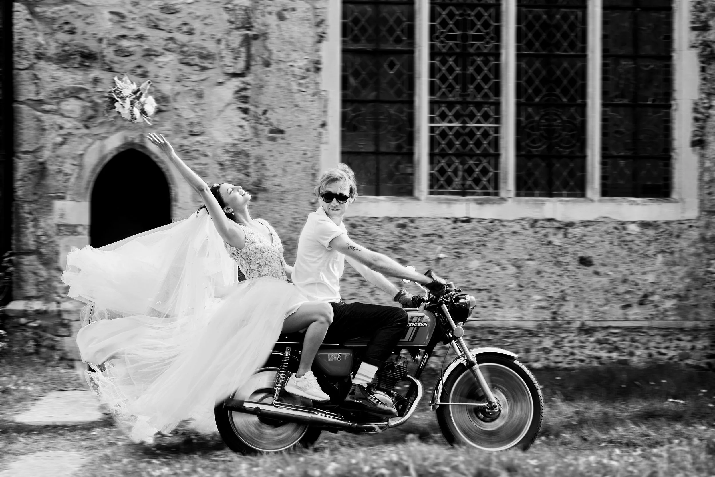 A person wearing sunglasses riding a motorcycle with a woman in a wedding dress sitting behind them, her arm raised and a bouquet thrown in the air. They are passing by an old stone building with large windows.