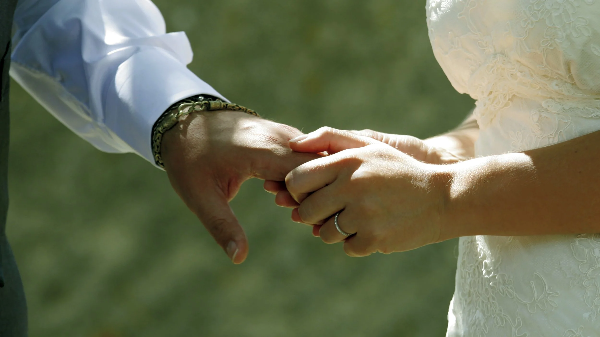Close-up of a man and woman holding hands, with the man's hand on top. The man is wearing a white long-sleeve shirt with jewelry on his wrist, and the woman has a ring on her finger and a cream-colored lace dress.