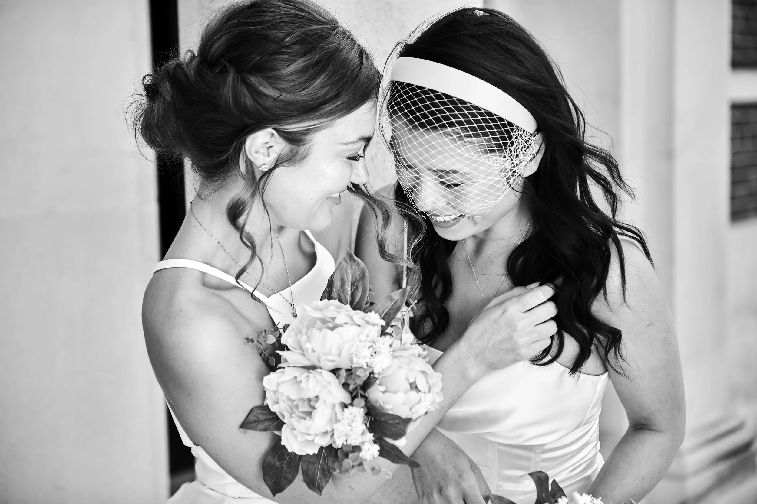 Two women in wedding dresses sharing a joyful moment, one holding a bouquet of flowers, black and white photograph.