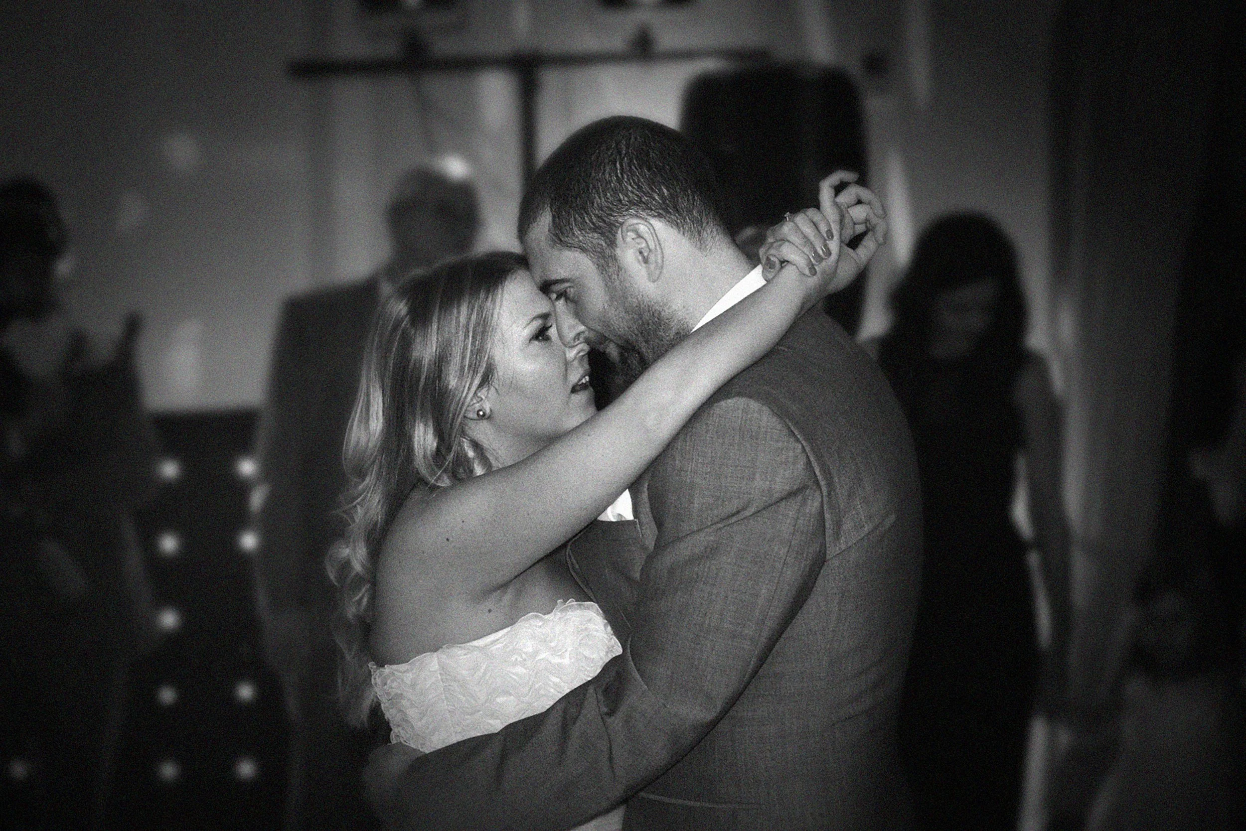 A black-and-white photo of a bride and groom dancing closely during their wedding reception, looking into each other's eyes. The bride has long, wavy hair and is wearing an off-the-shoulder dress, while the groom has short hair and a beard, dressed i