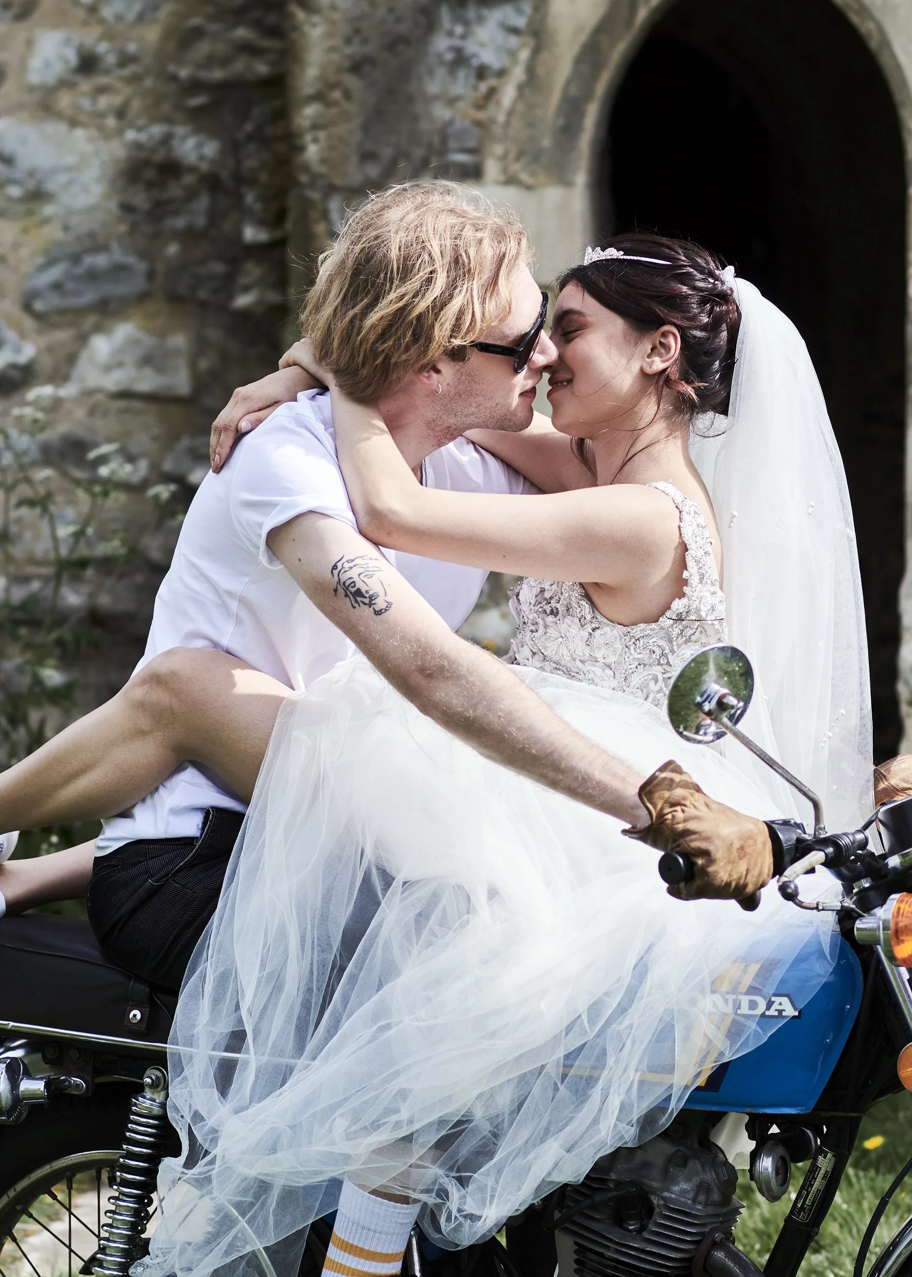 A newlywed couple, dressed in wedding attire, sharing a kiss while riding a vintage Honda motorcycle outdoors near a stone building.