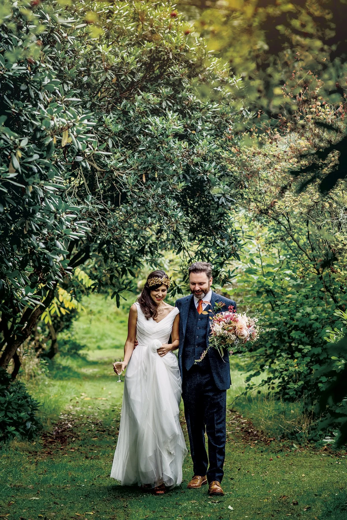 A bride and groom walking together on a grassy path surrounded by lush green trees, with the groom holding a bouquet of flowers.