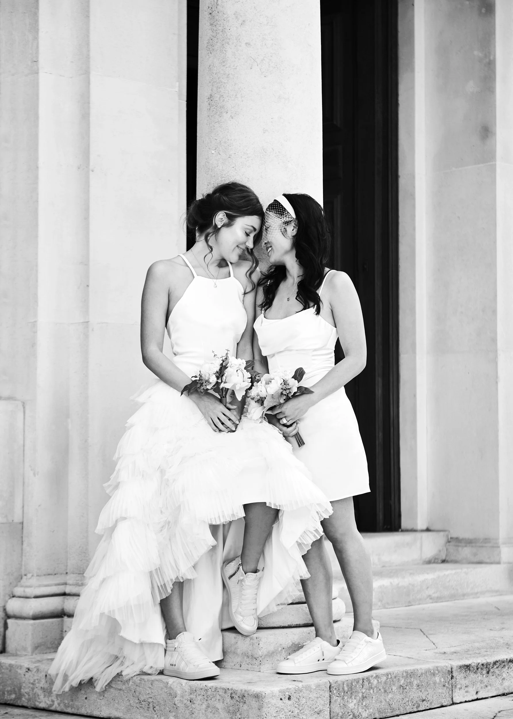 Two women in wedding dresses smiling and touching foreheads while holding bouquets, standing on the steps outside a building.