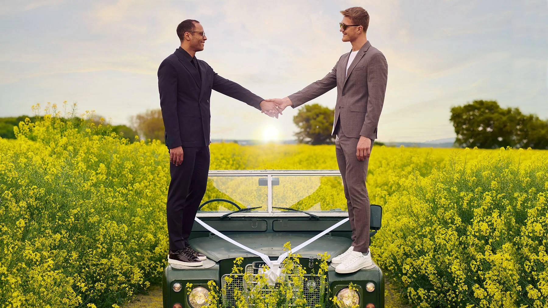 Two men in business suits, one older and one younger, shaking hands while standing on the hood of a vintage vehicle in a yellow flowering field during sunset.