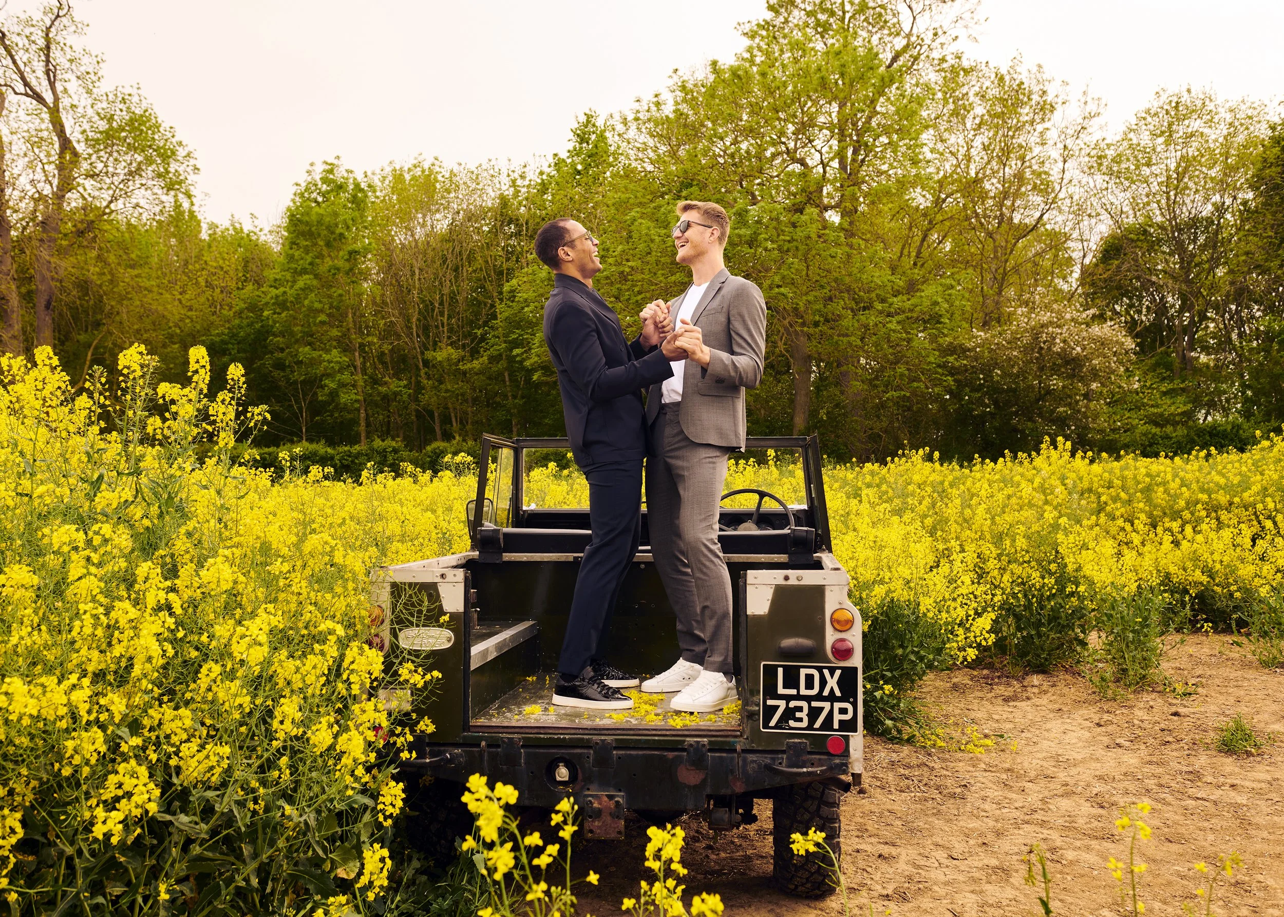 Two men in suits celebrating and holding hands on the back of a small vehicle in a yellow flower field, with green trees in the background.