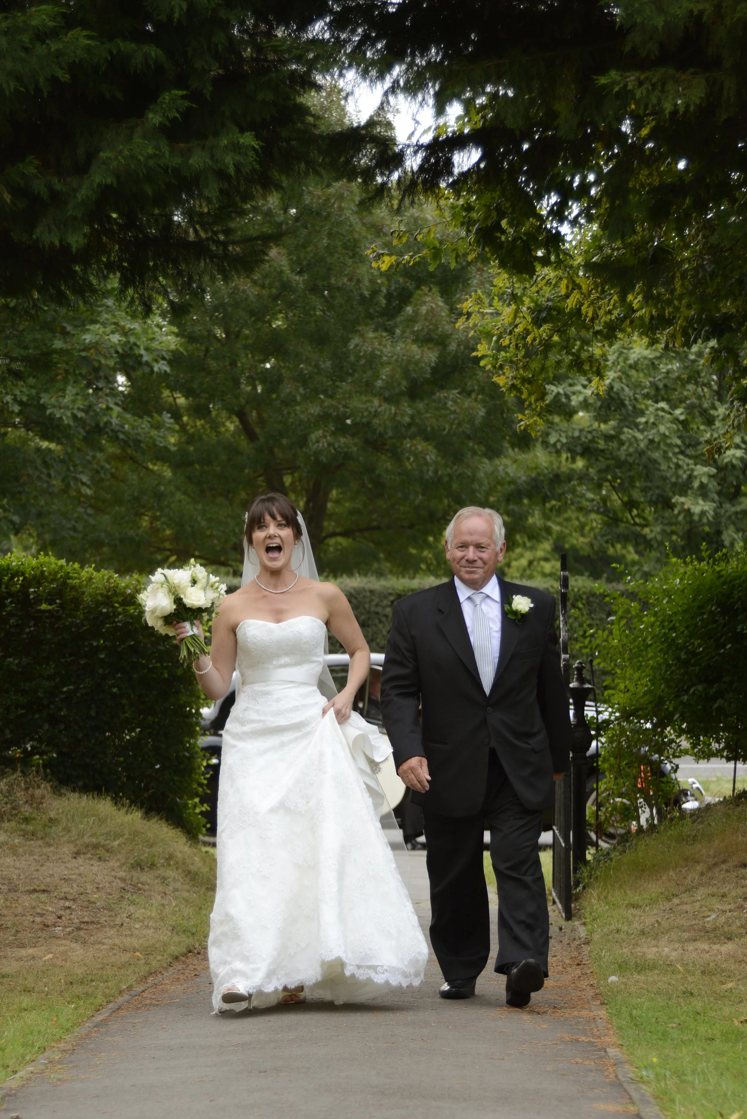 A bride in a white wedding dress holding a bouquet, smiling and walking arm-in-arm with an older man in a black suit, outdoors on a paved path surrounded by greenery.