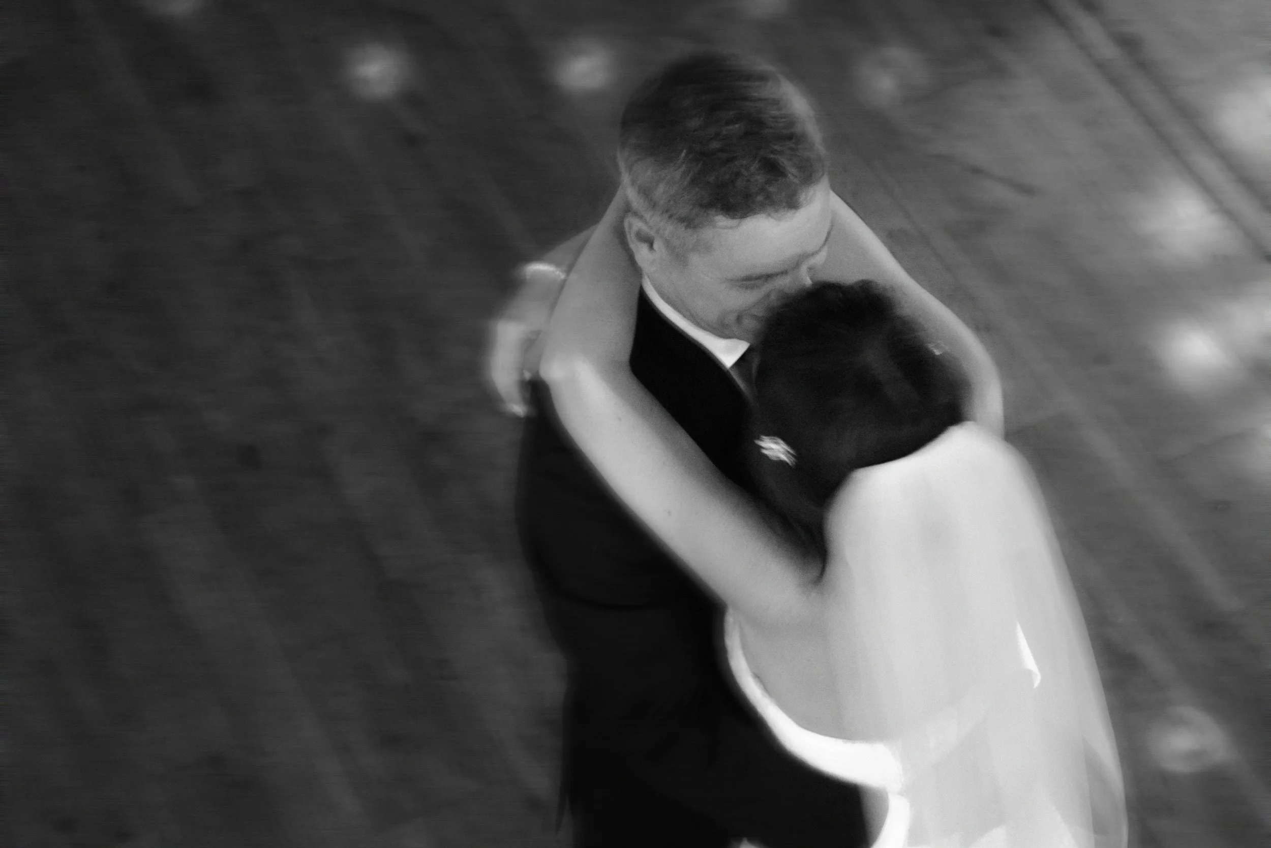 A black and white aerial photograph of a couple dancing, with the man in a tuxedo and the woman in a light-colored dress, embracing on a wooden dance floor.
