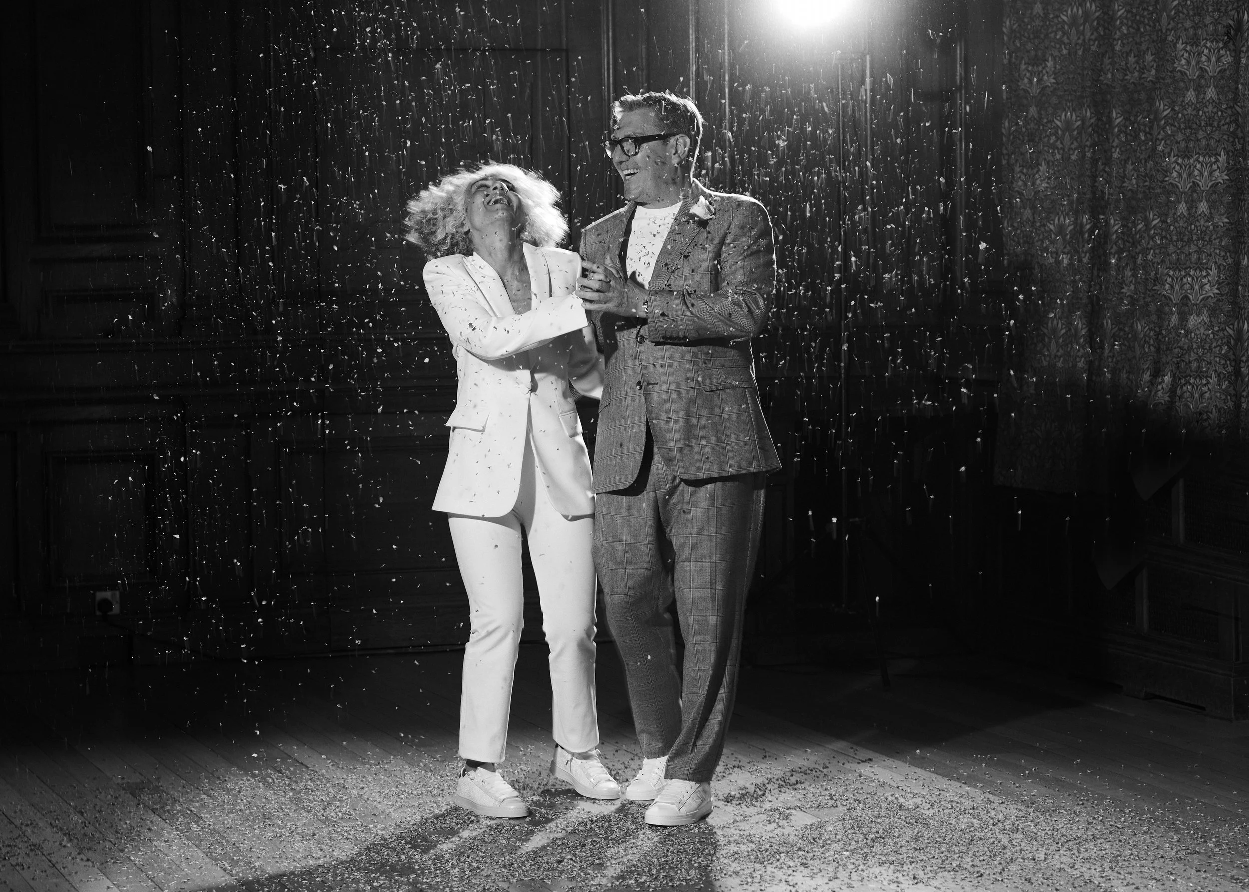 Two people, a woman and a man, smiling and celebrating while dancing together, with confetti falling around them in a room with wood-paneled walls and patterned wallpaper.