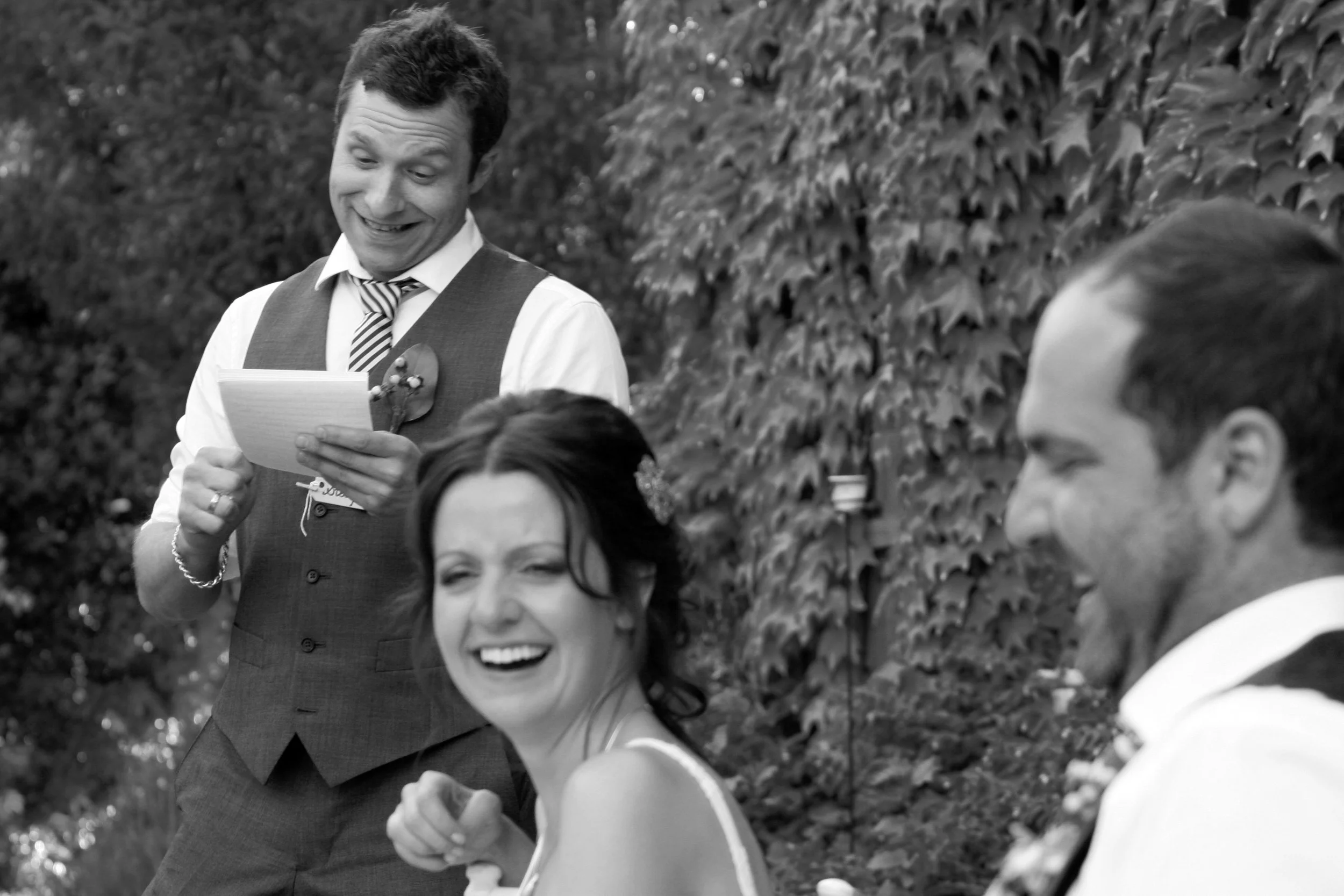 Black and white photo of a bride and groom with a man reading a speech at an outdoor wedding reception, surrounded by leafy vines.