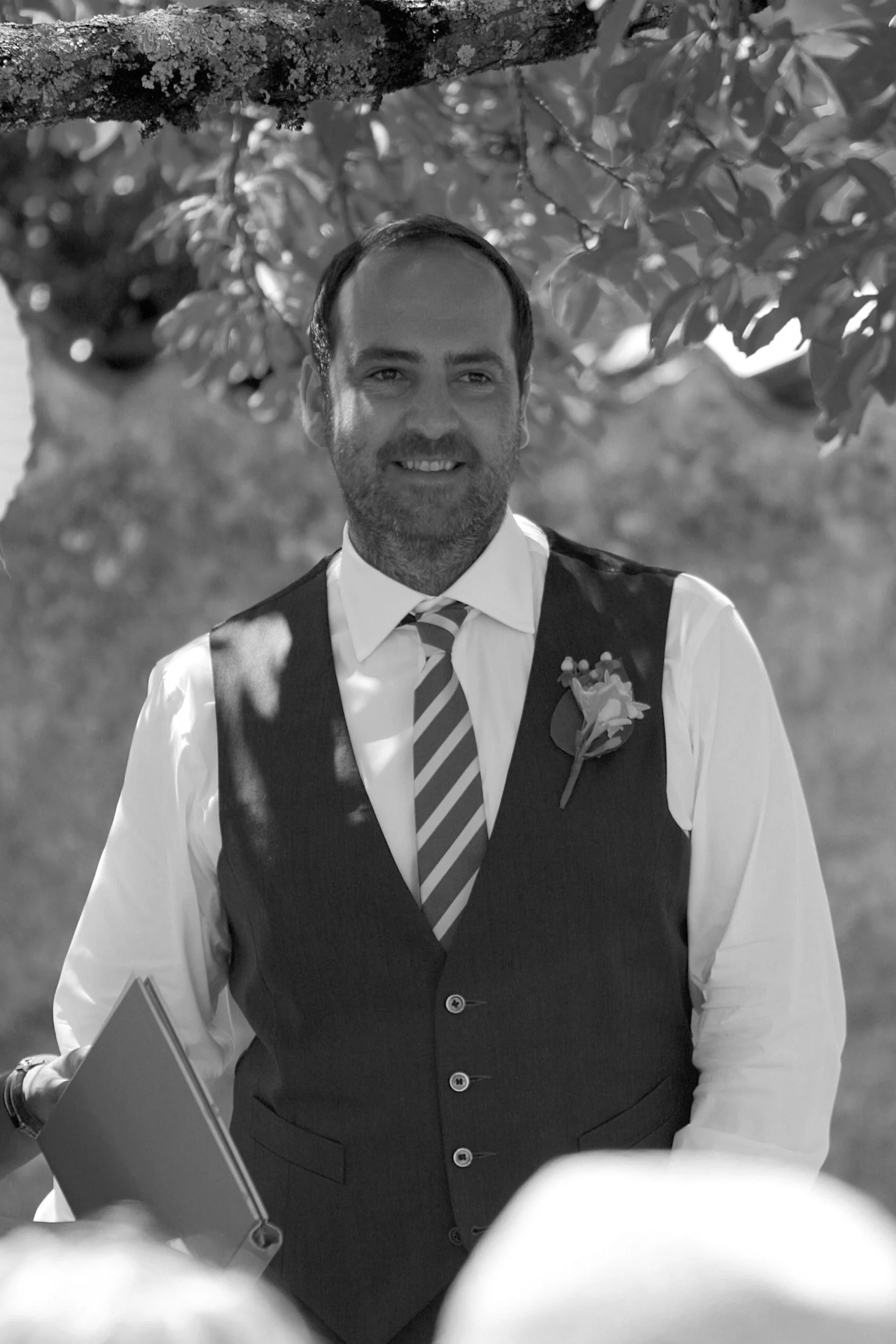 A man smiling outdoors, wearing a white shirt, striped tie, dark vest with a flower boutonniere, holding a folder, under a tree with leaves and branches.