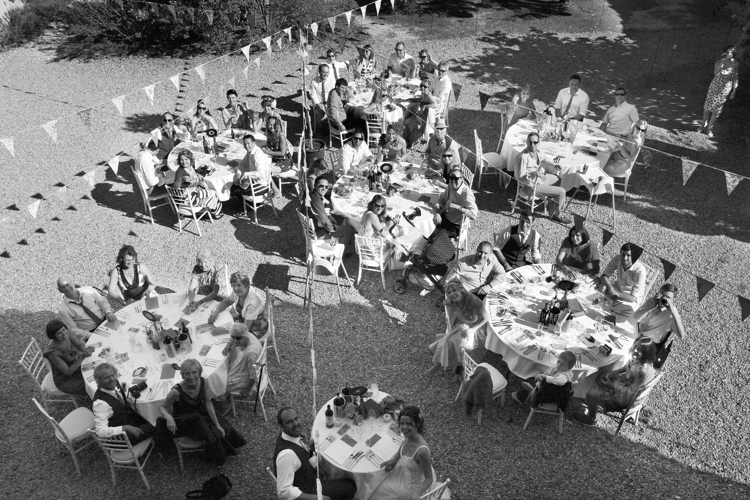Black and white photo of an outdoor celebration with multiple round tables filled with people, set on a gravel surface. People are eating, drinking, and engaging in conversation. Bunting flags hang overhead, and shadows are cast from nearby trees.