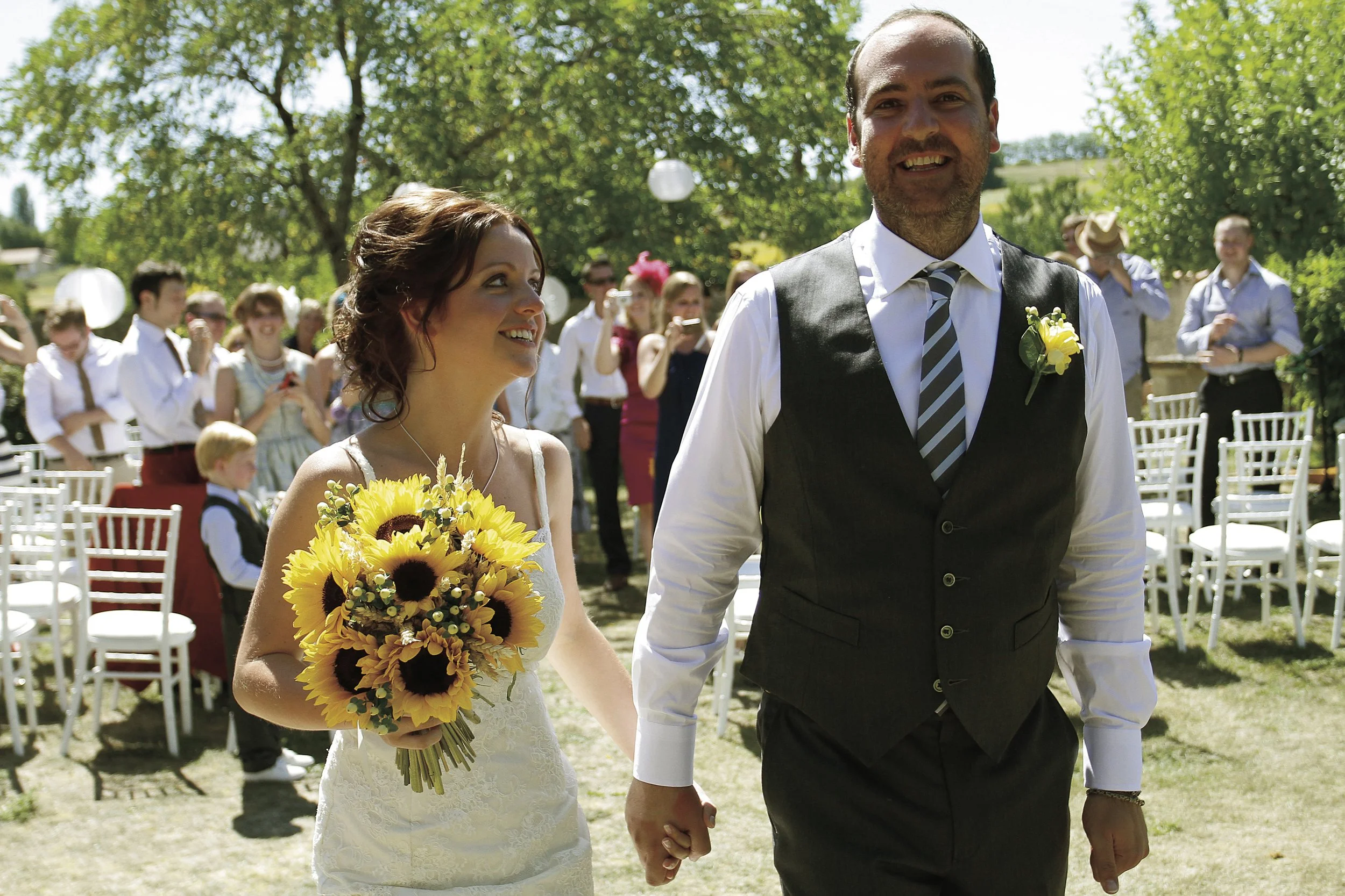 A bride and groom walking hand in hand at their outdoor wedding ceremony, surrounded by guests and greenery.
