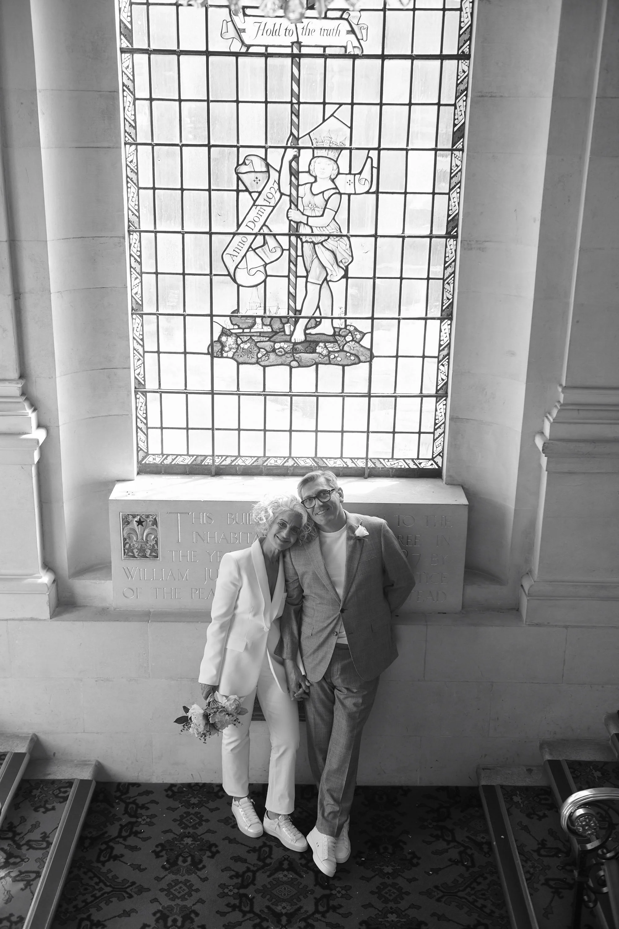 A black-and-white photo of a smiling couple standing close together in front of a large stained glass window showing a soldier holding a flag, inside a building with decorative stone walls and a patterned carpet.