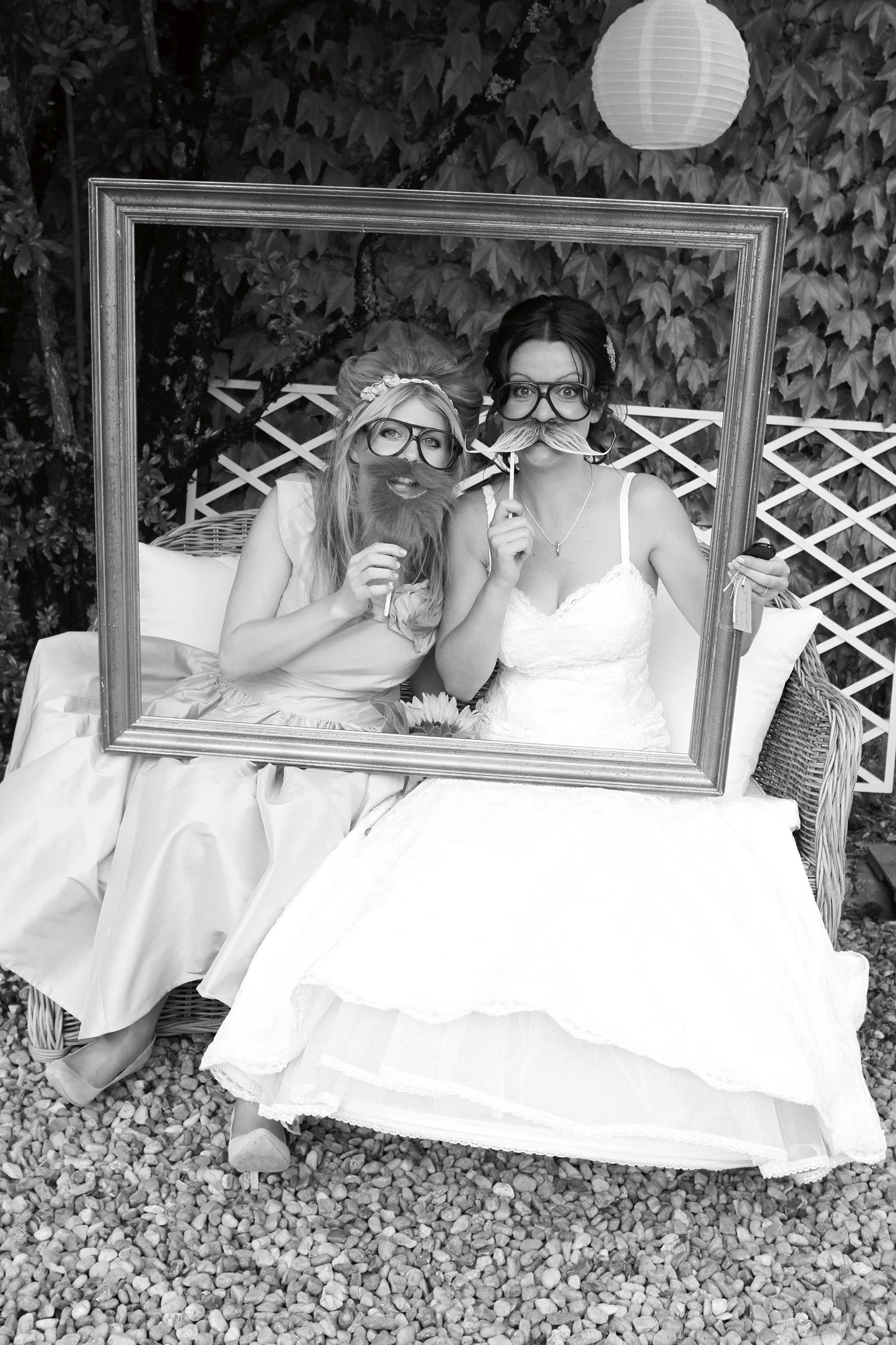 Two women sitting on a garden bench, wearing wedding dresses, holding photo booth props with glasses and beards, inside a large picture frame, outdoor setting with gravel ground and leafy background.