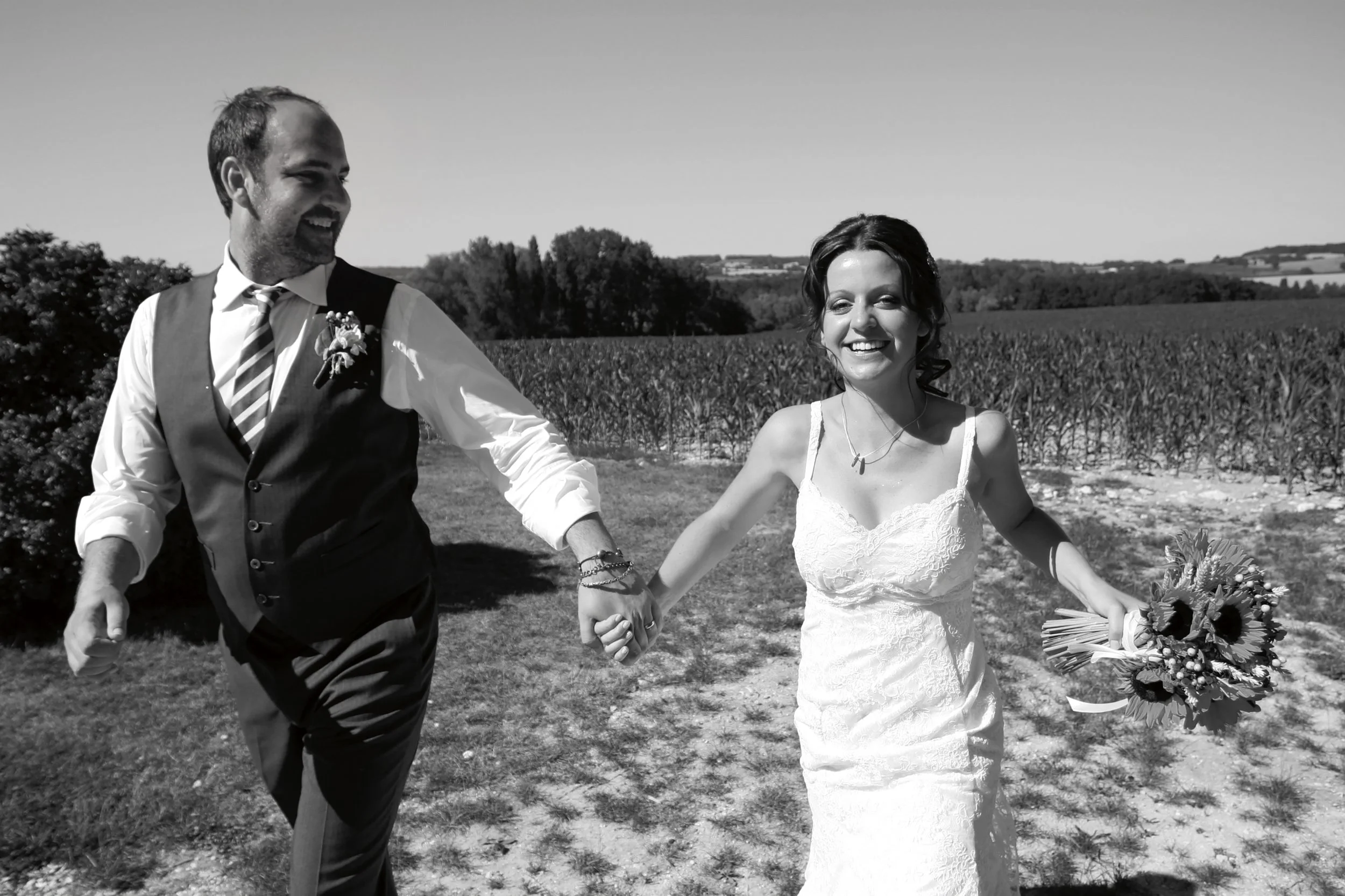 A smiling bride and groom holding hands outdoors in a rural landscape, with a bouquet of sunflowers in the bride's hand.