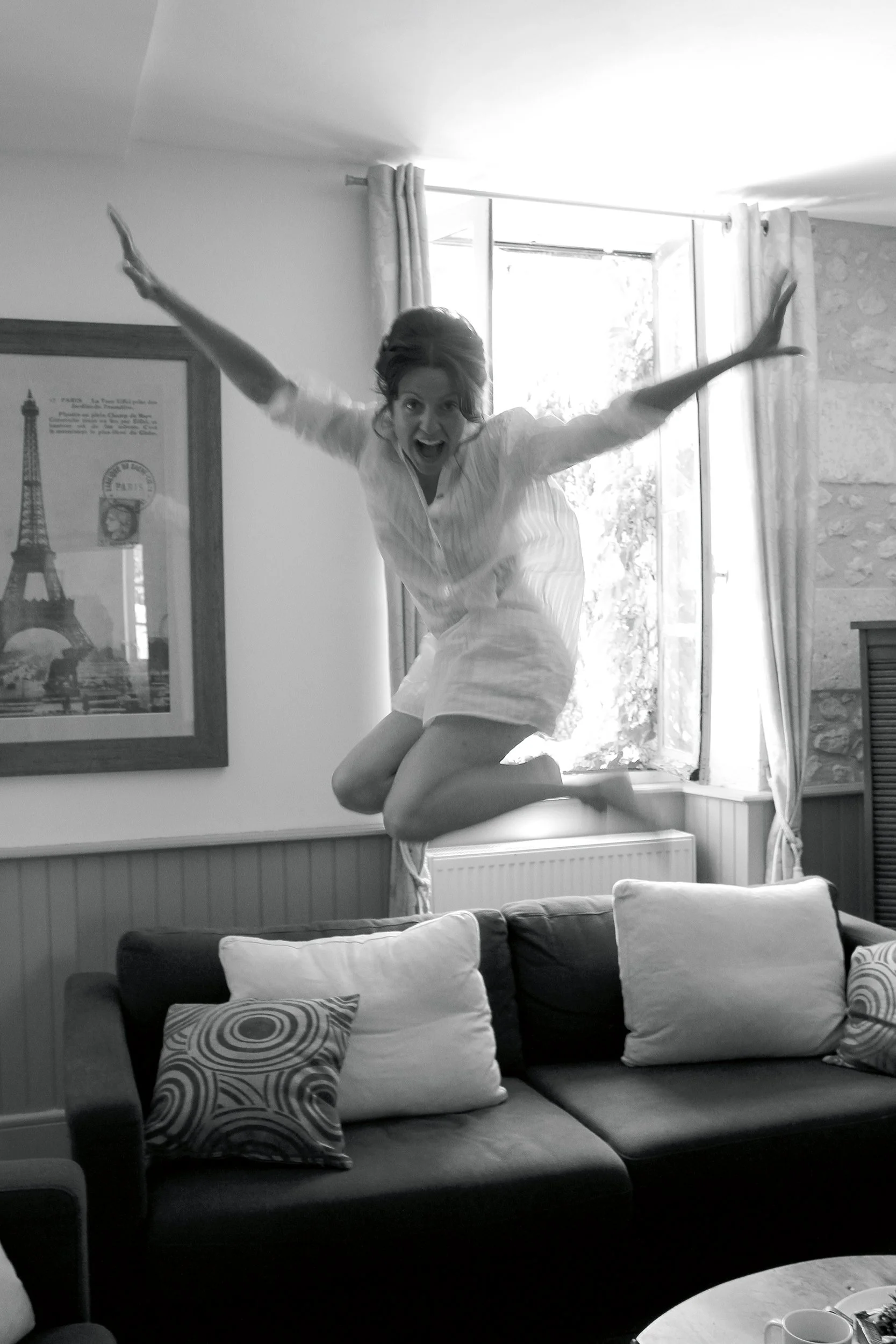 A woman jumping on a couch in a living room, smiling with arms outstretched, black and white photo.