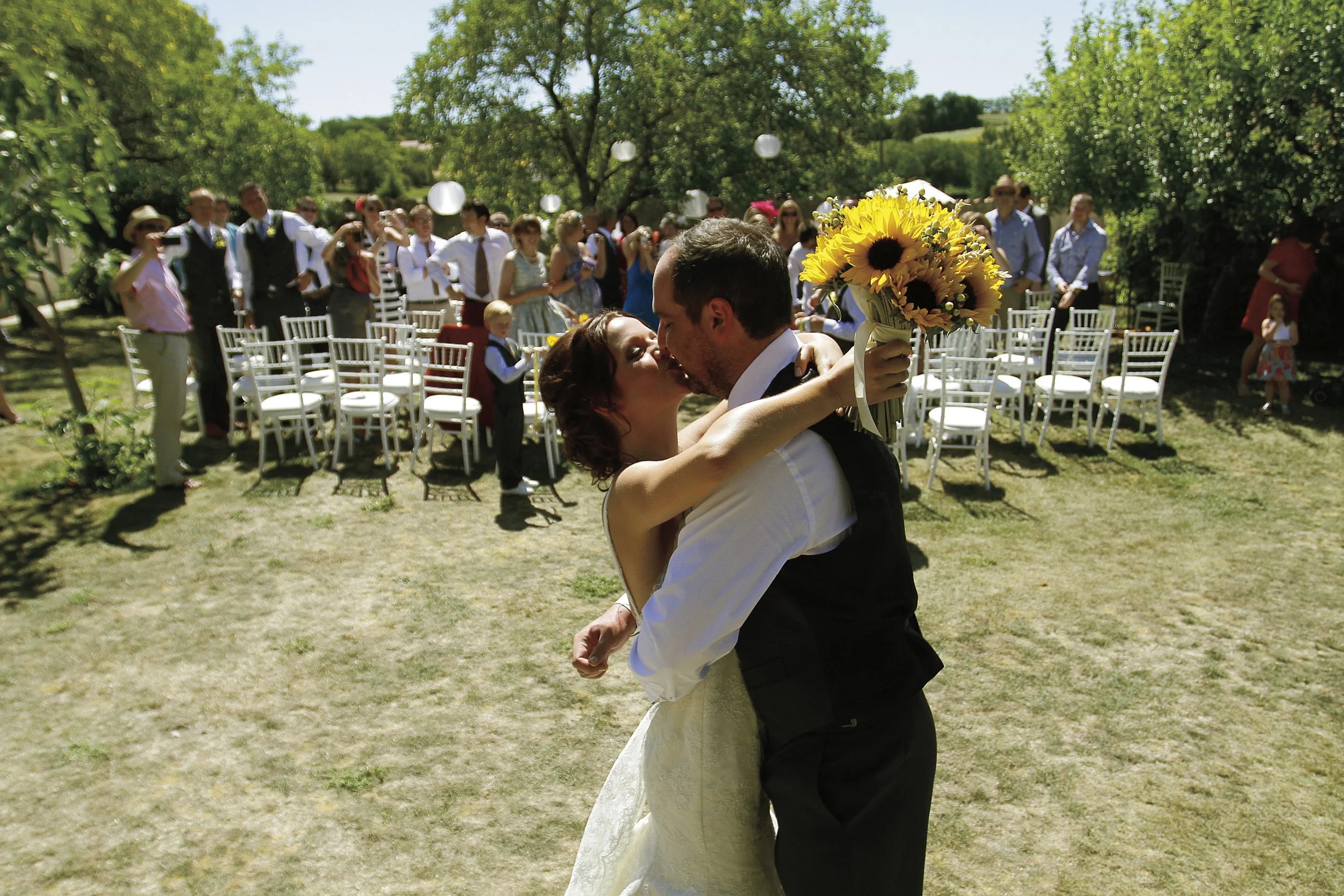 A bride and groom kissing outdoors during their wedding with guests seated and standing in the background on a sunny day.