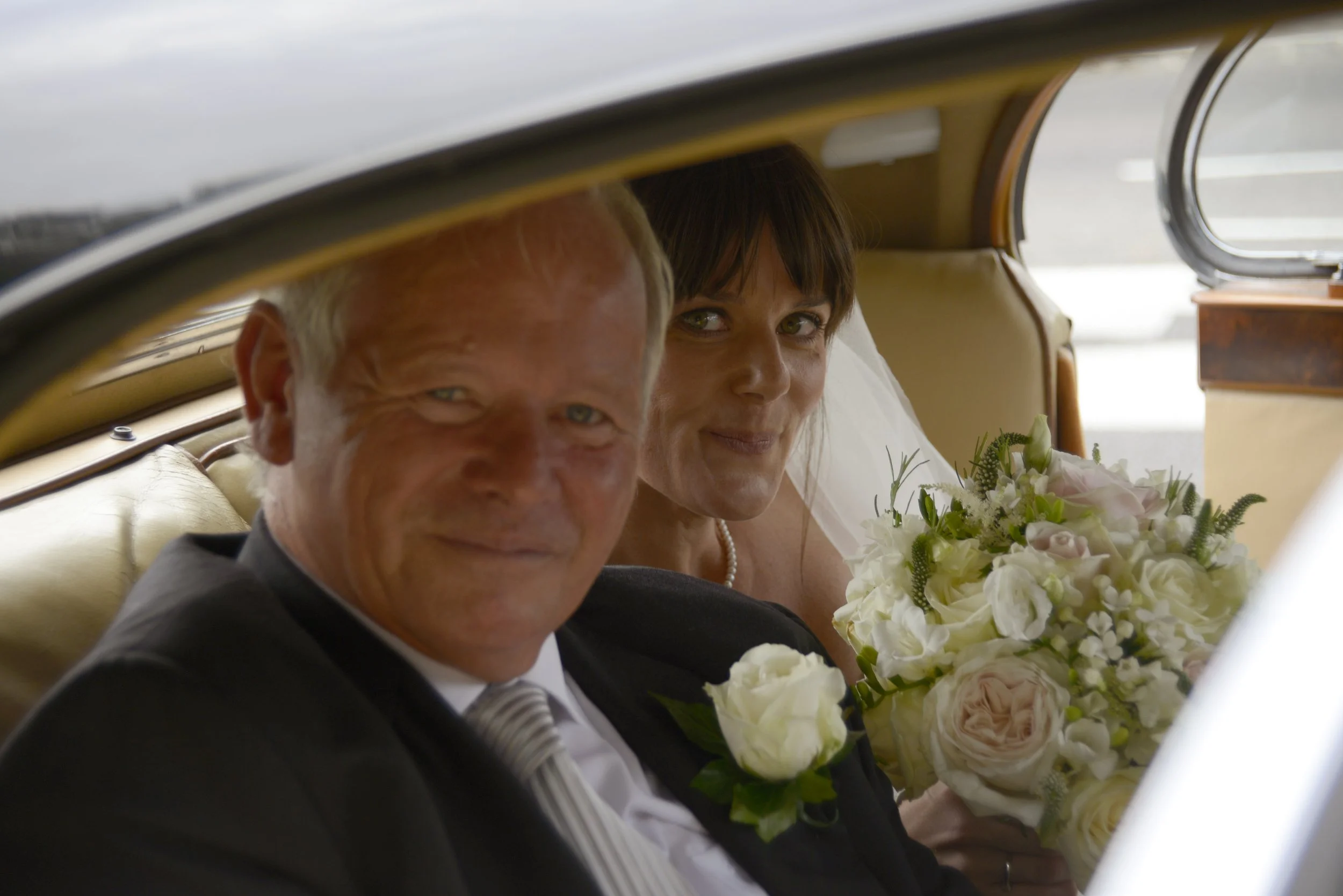 A bride and groom sitting inside a vintage car, smiling at the camera. The bride holds a bouquet of white and pale pink flowers.