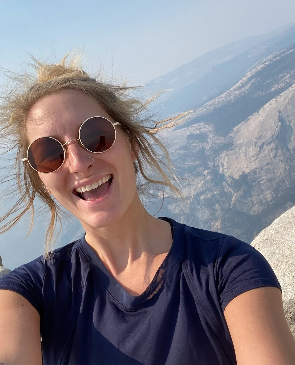 A woman with sunglasses and a nose ring taking a selfie on a mountain summit, with a rocky mountain range in the background.
