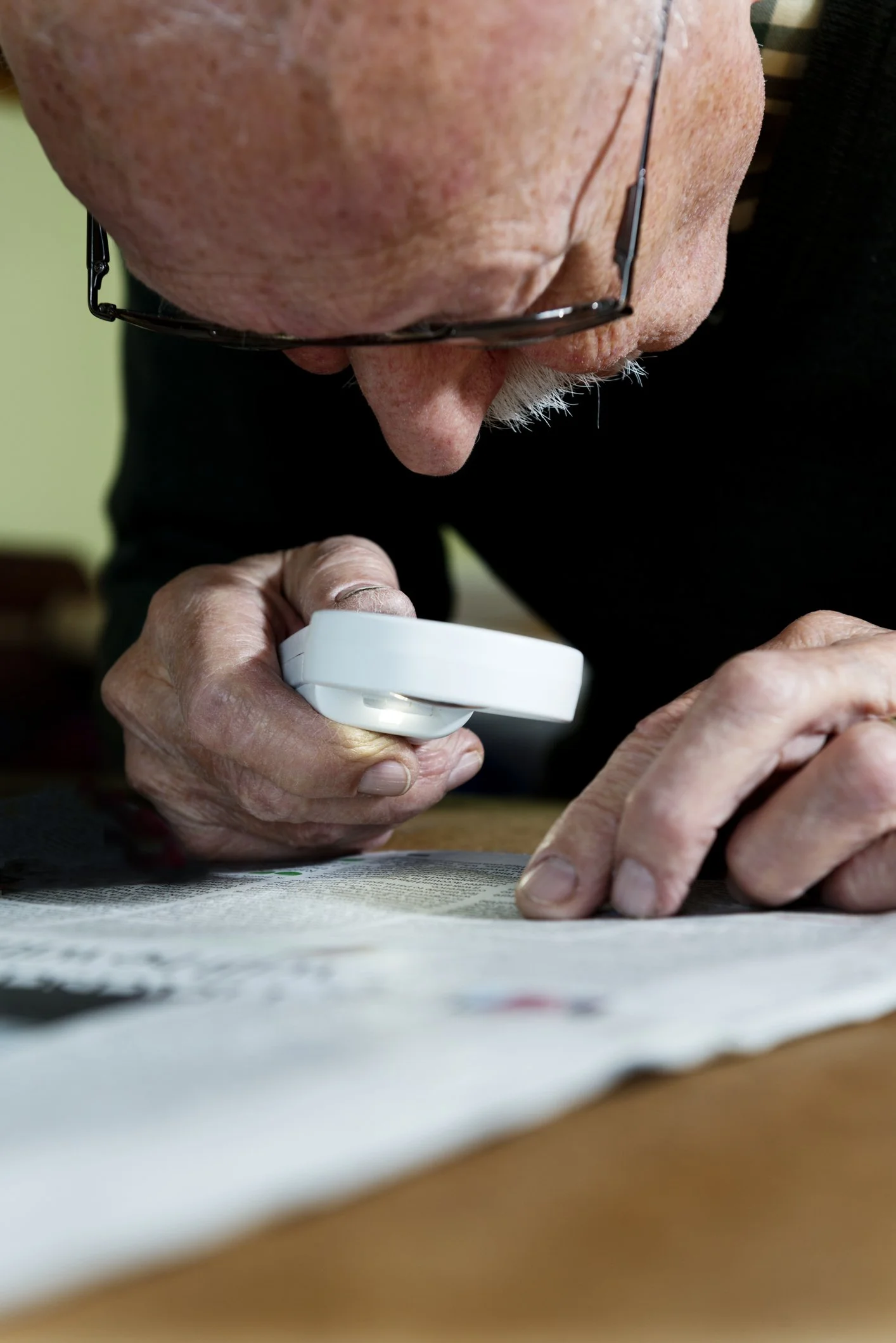 Close-up of an elderly man with glasses using a magnifying glass to closely examine a newspaper on a table.