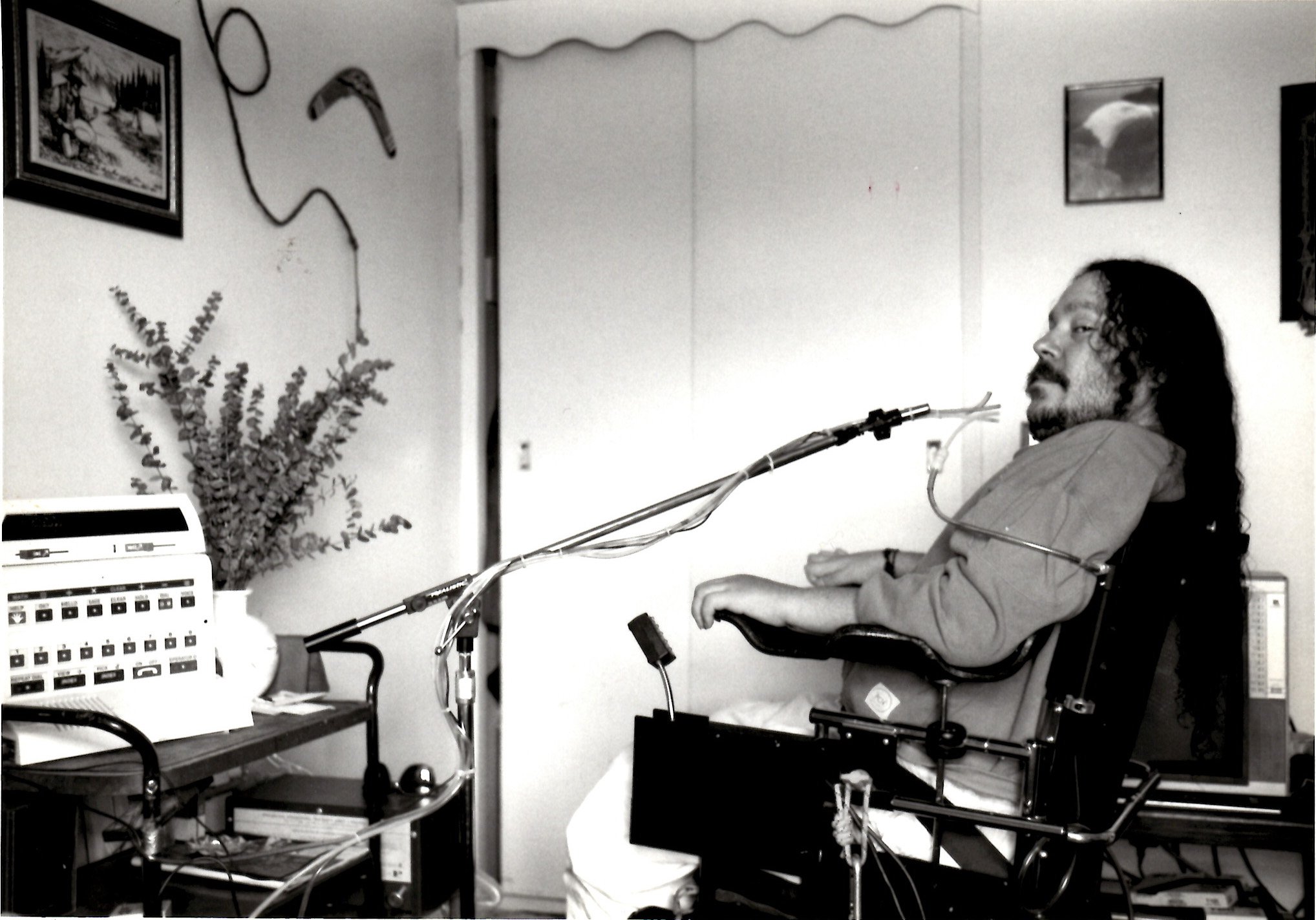 A man with long hair and a beard sitting in a wheelchair with medical equipment attached, in a room with framed pictures and a cabinet with electronic devices.