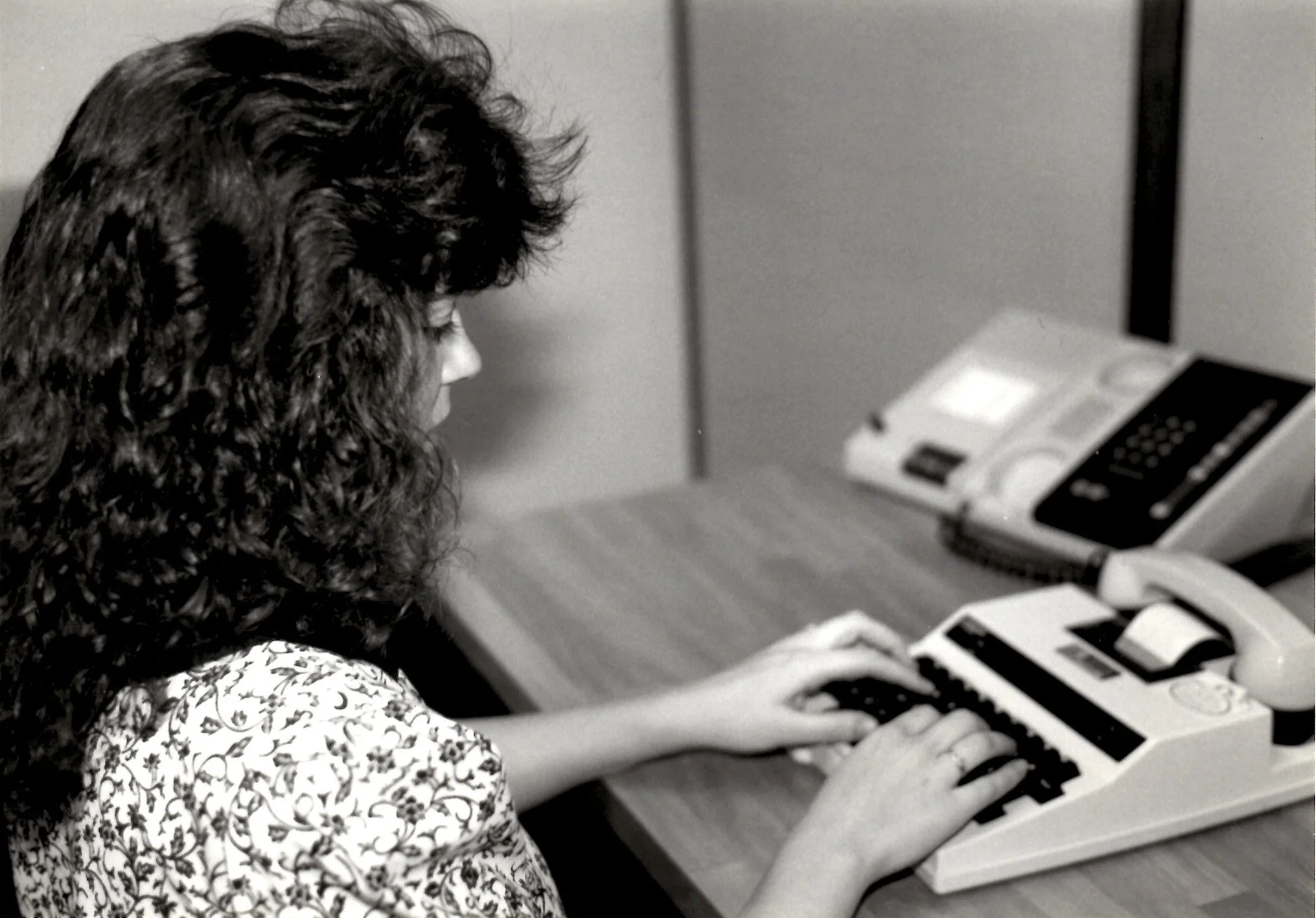 A woman with curly hair using a vintage desktop calculator in an office environment.