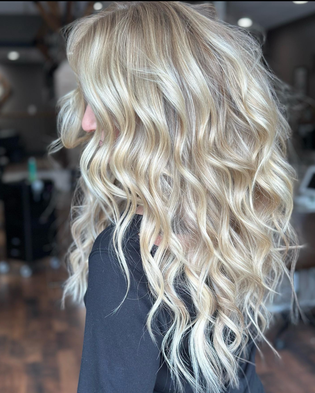 Side view of a woman with long, blonde, wavy hair. She is in a salon or hair studio with blurred background showing salon equipment.