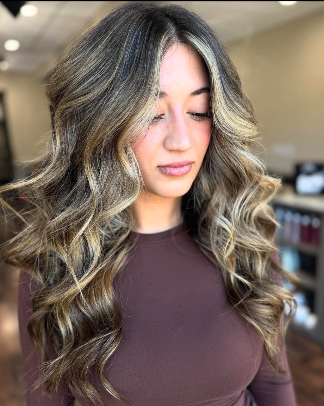 A young woman with long, wavy, highlighted hair and light makeup, standing indoors with a blurred background.